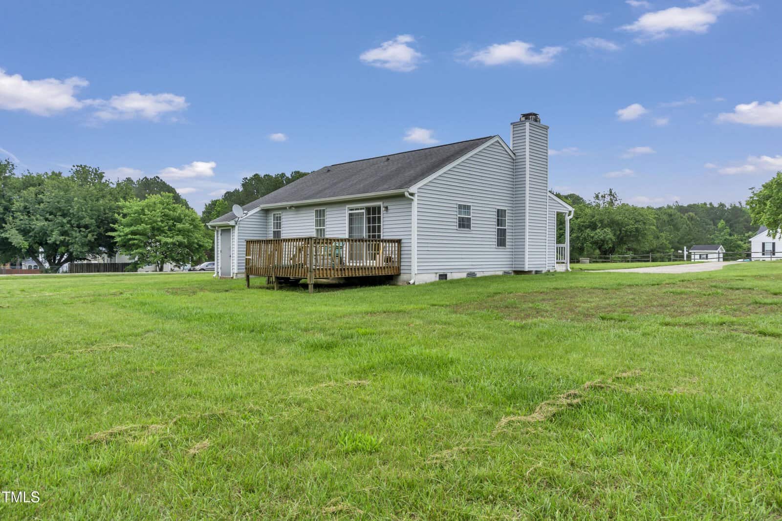 Single-story white house with a wooden deck in the backyard, surrounded by a large grassy lawn and trees. The sky is partly cloudy, and a driveway is visible on the right side.