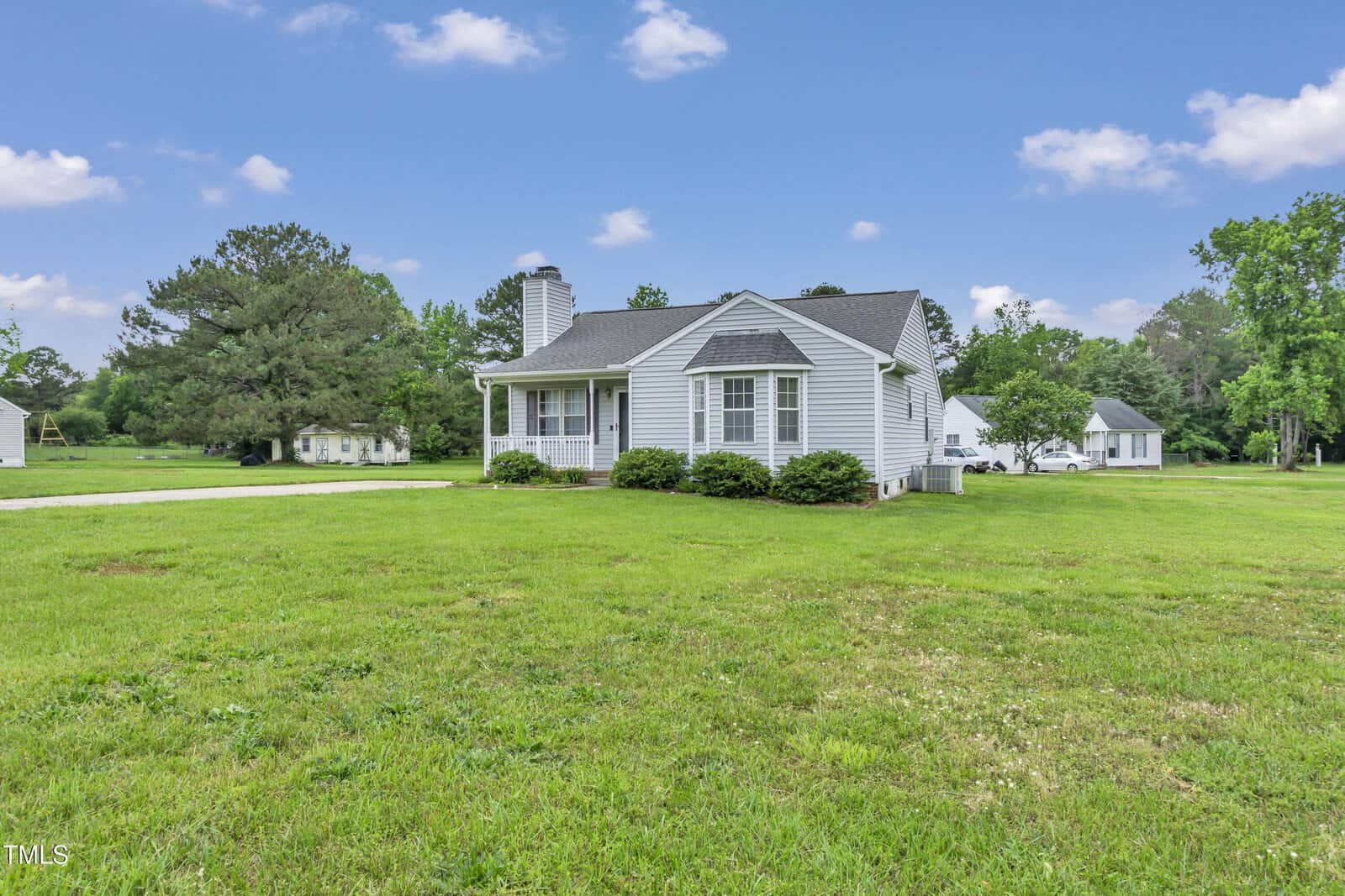 A small, single-story gray house with white trim sits on a large, grassy lawn with a few bushes in front. Trees and another house are visible in the background under a partly cloudy sky.