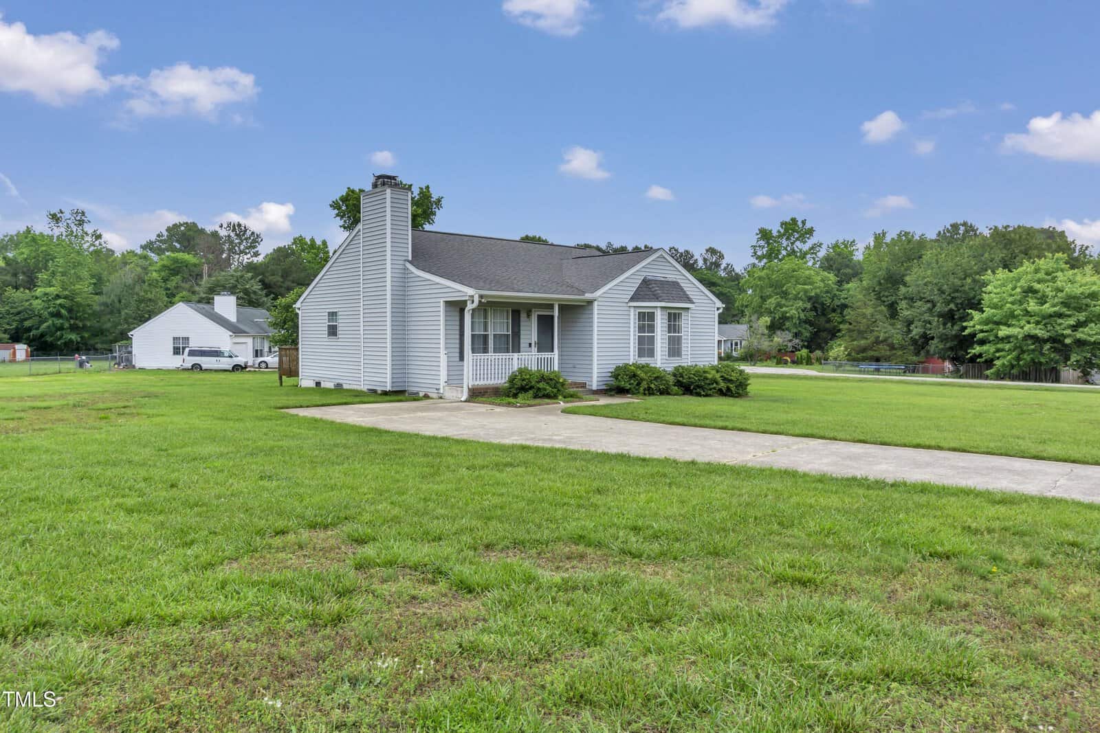 A small, single-story gray house with white trim sits on a large grassy lot with a concrete driveway. Trees and another house are visible in the background under a blue sky with scattered clouds.