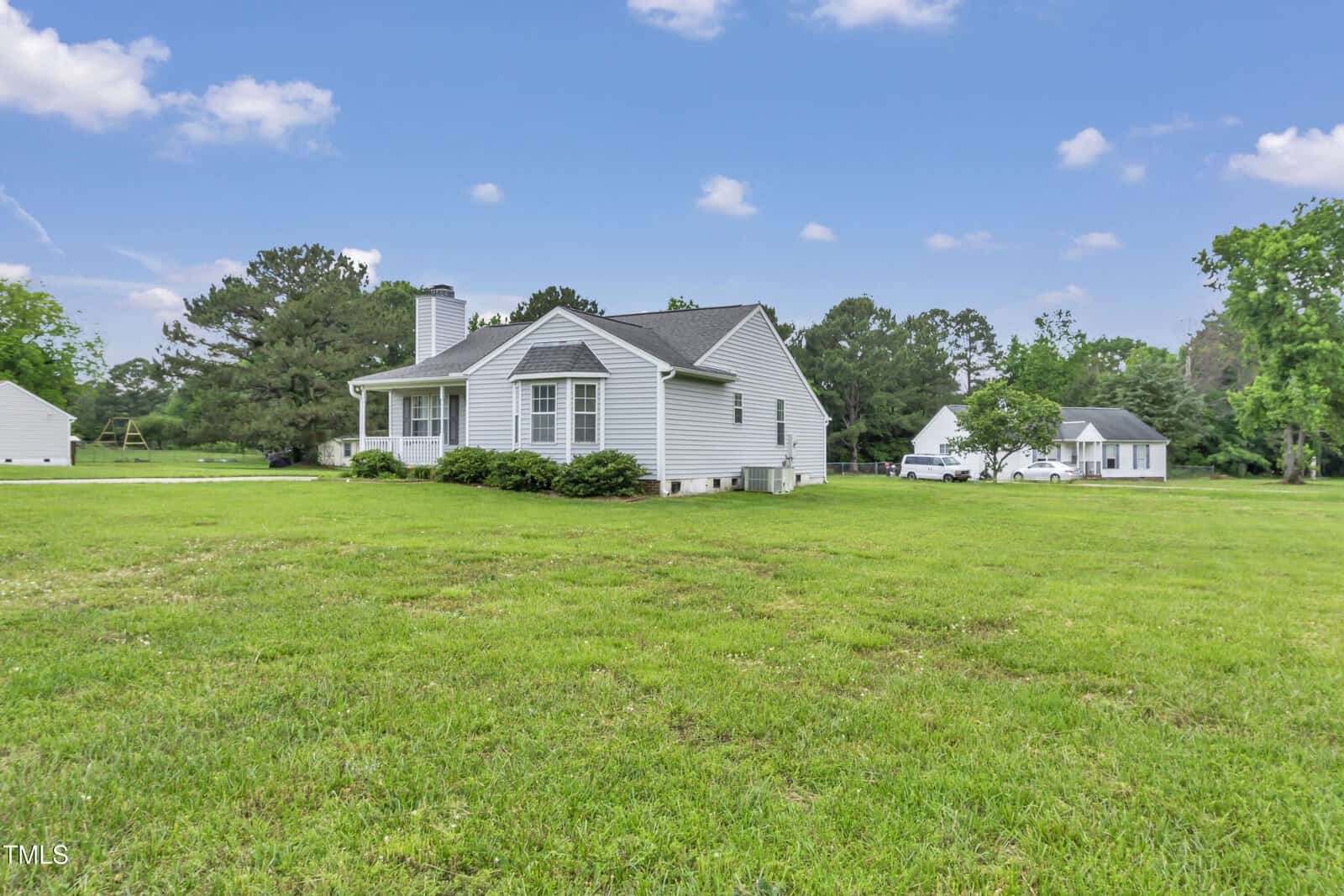 A small gray house with a front porch sits on a large, grassy lawn with trees in the background and a detached white garage or outbuilding nearby under a blue sky with scattered clouds.