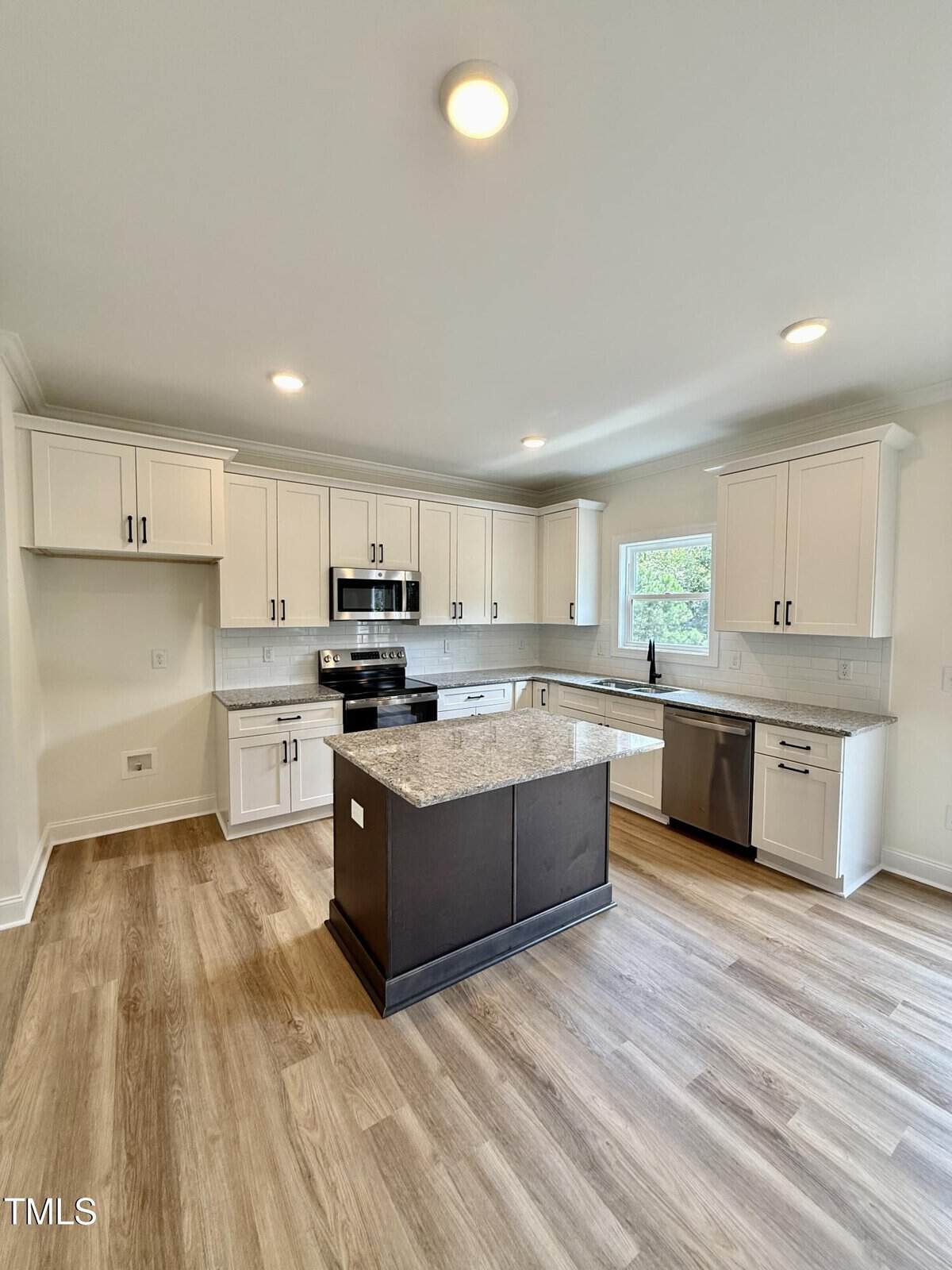 Modern kitchen with white cabinets, stainless steel appliances, a center island with granite countertop, light wood flooring, and a window above the sink allowing natural light in.