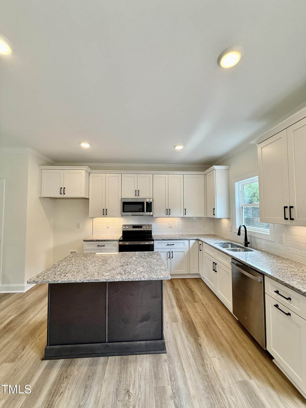 Modern kitchen with white cabinets, stainless steel appliances, granite countertops, a large center island with dark base, light wood flooring, and recessed lighting. A window above the sink lets in natural light.