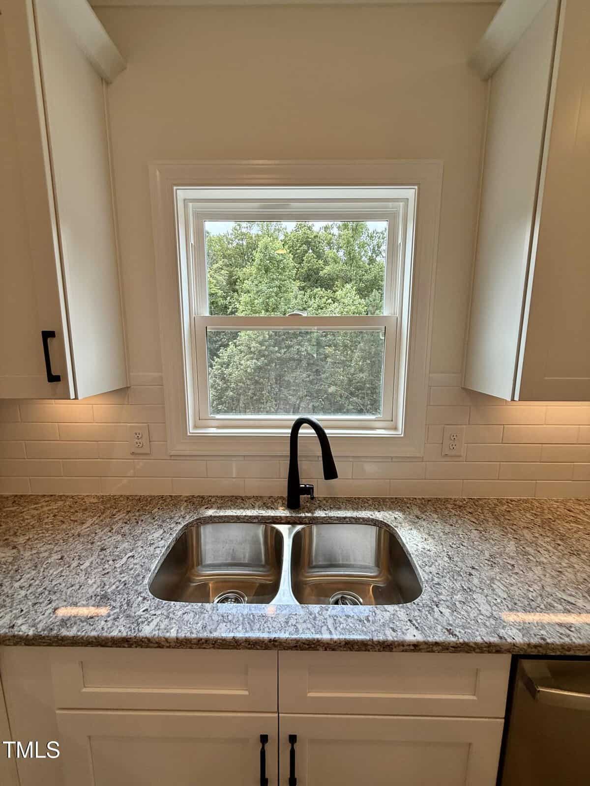 A kitchen sink with a black faucet is set in a granite countertop beneath a window, which overlooks green trees. White cabinets and subway tile backsplash surround the area.