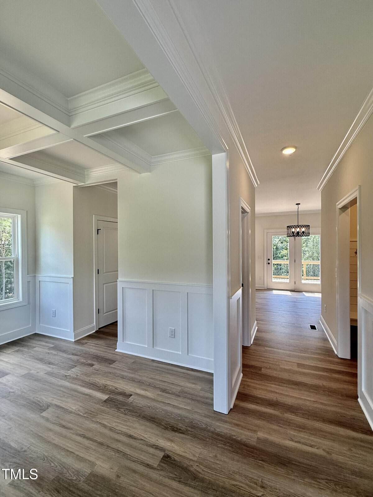 A bright hallway with white walls, coffered ceiling, wood flooring, and doorways leading to rooms. Sunlight streams through windows and glass doors at the far end, creating a welcoming atmosphere.