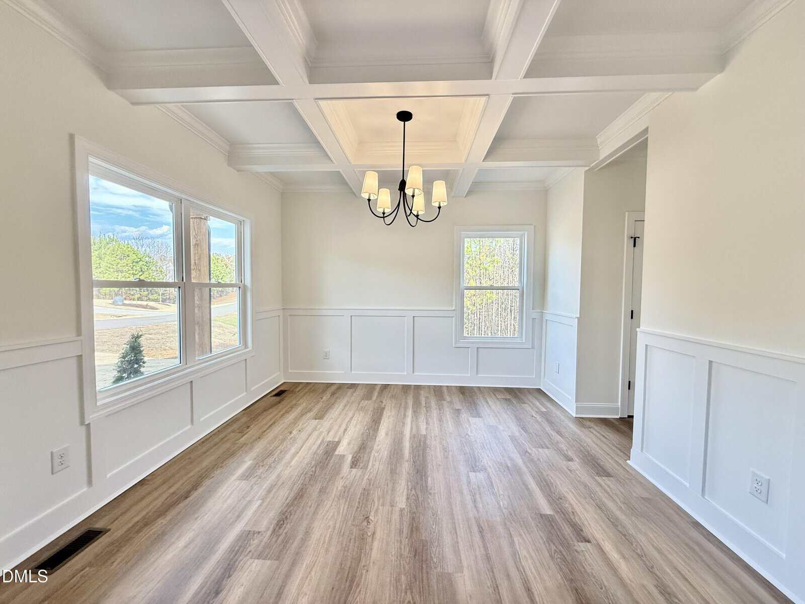 Bright dining room with large windows, coffered ceiling, chandelier, white wainscoting, and light wood flooring. Outdoor scenery is visible through the windows.