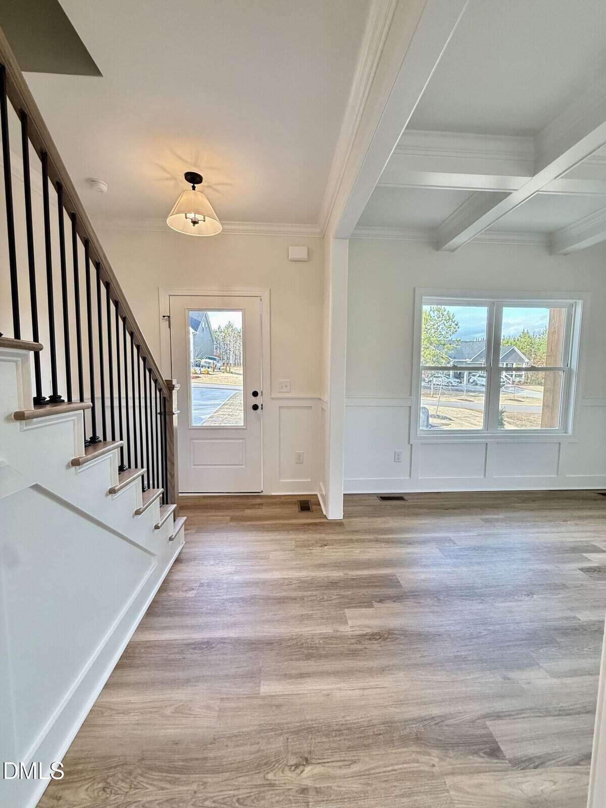 Bright entryway of a house with wooden stairs and black railing on the left, white walls, wood flooring, and large windows letting in natural light on the right. A light fixture hangs by the door.