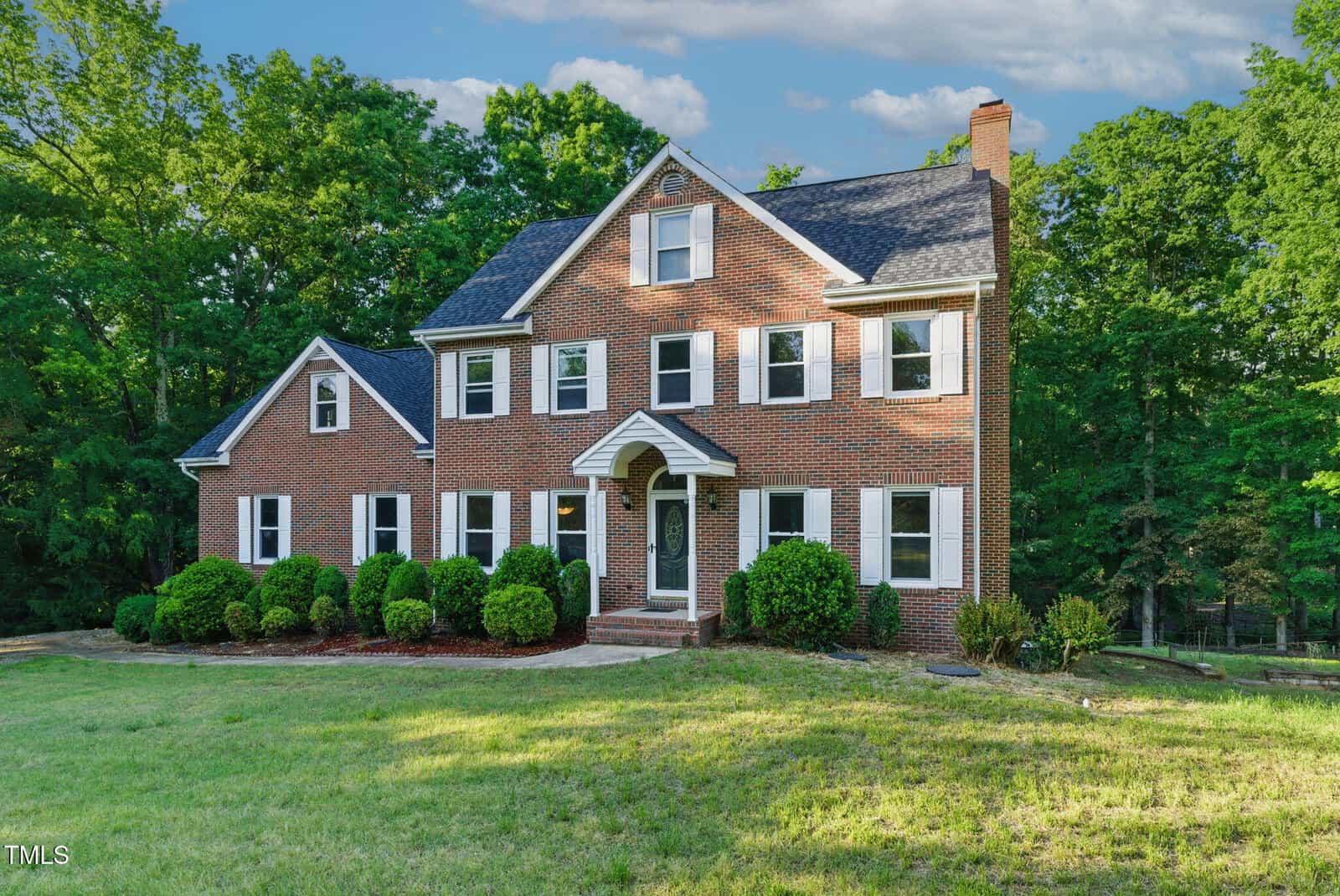 Three-story brick house with white shutters, arched front entry, and landscaped shrubs, set on a grassy lawn with trees in the background under a partly cloudy sky.