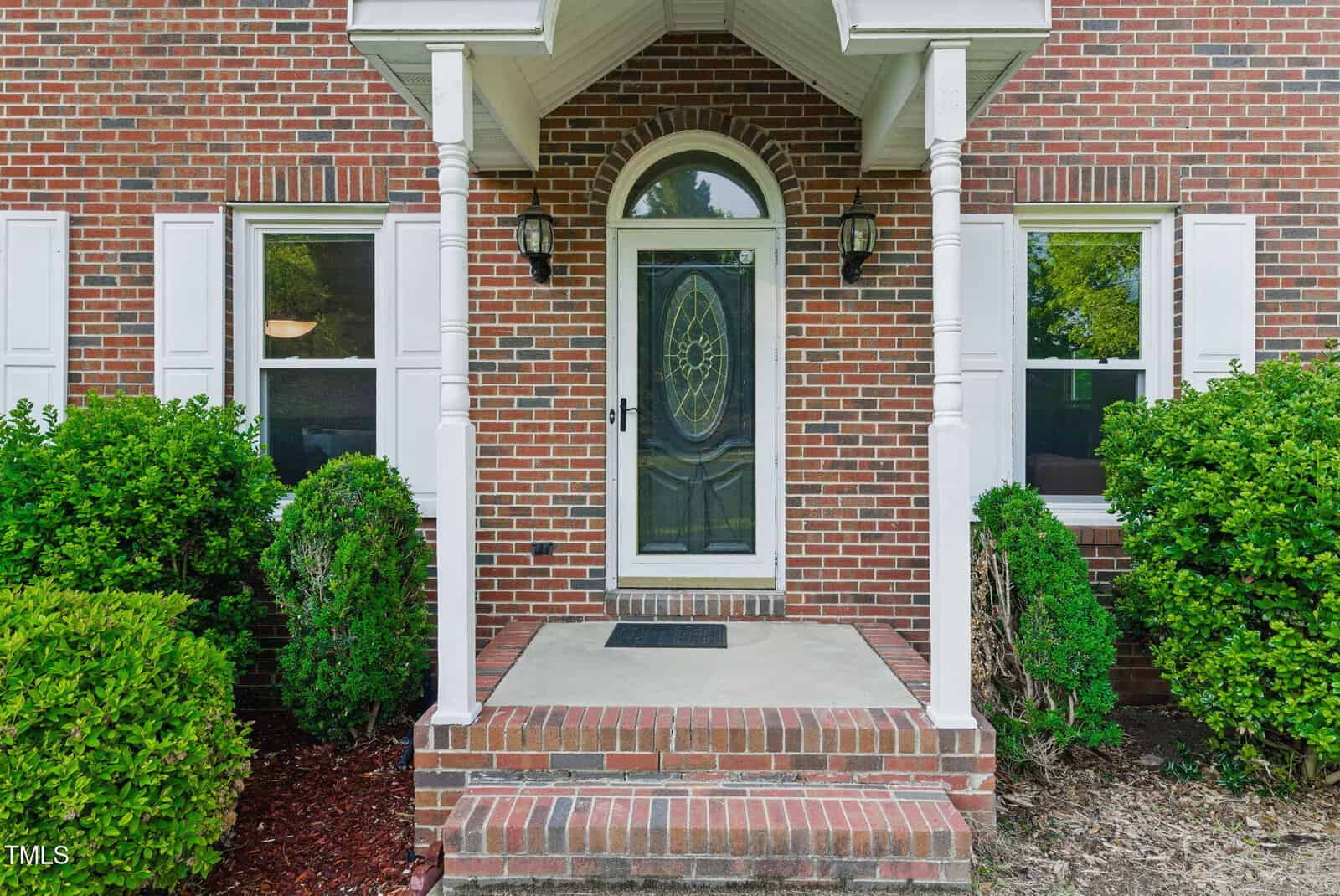 A brick house entryway with a black oval-glass front door, white trim, two white columns, two side windows with white shutters, and green bushes lining the small front porch.
