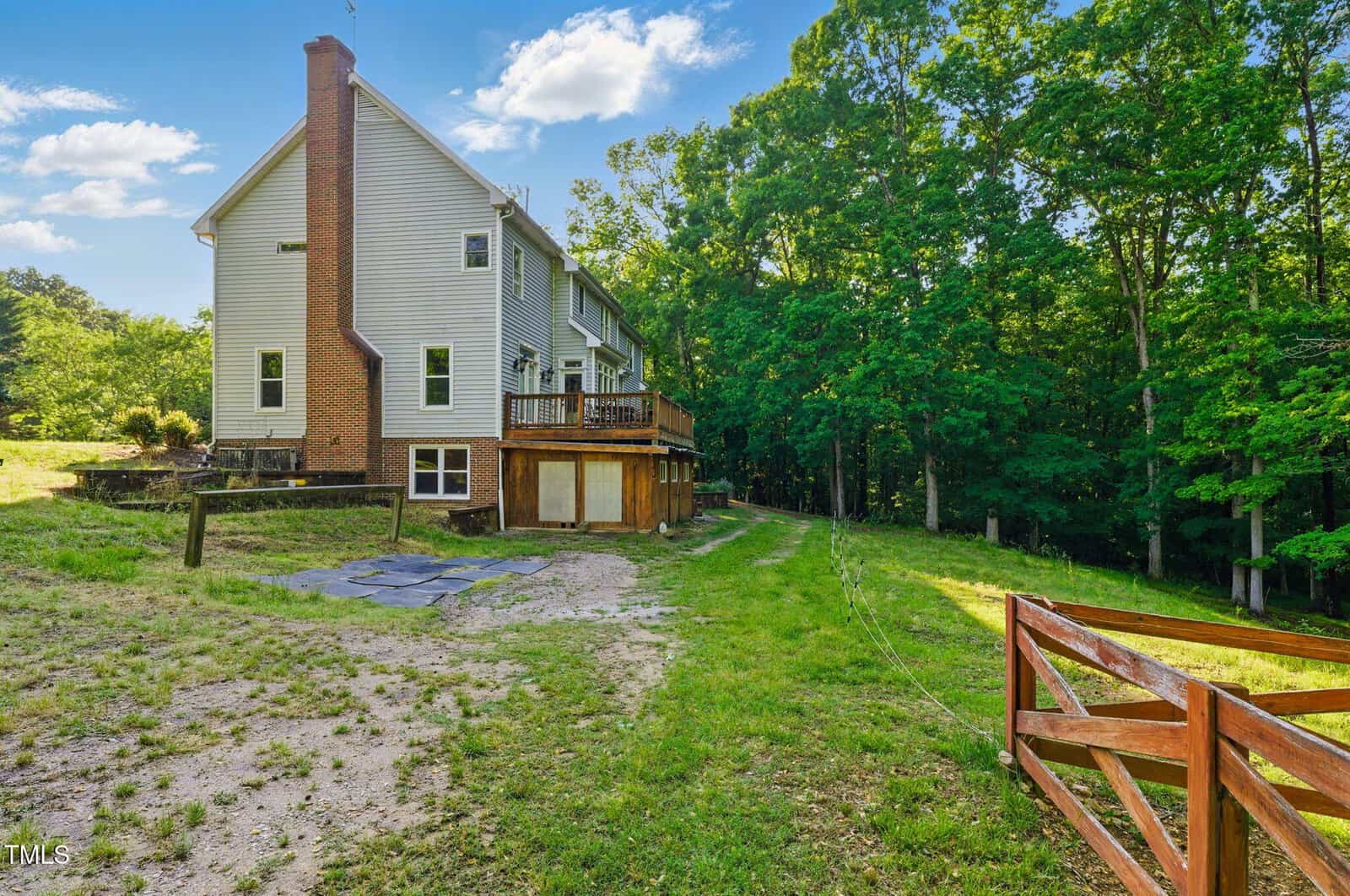A large two-story house with gray siding and a brick chimney sits next to a wooded area, featuring a wooden deck, a dirt path, and a red wooden fence in the foreground under a blue sky with scattered clouds.