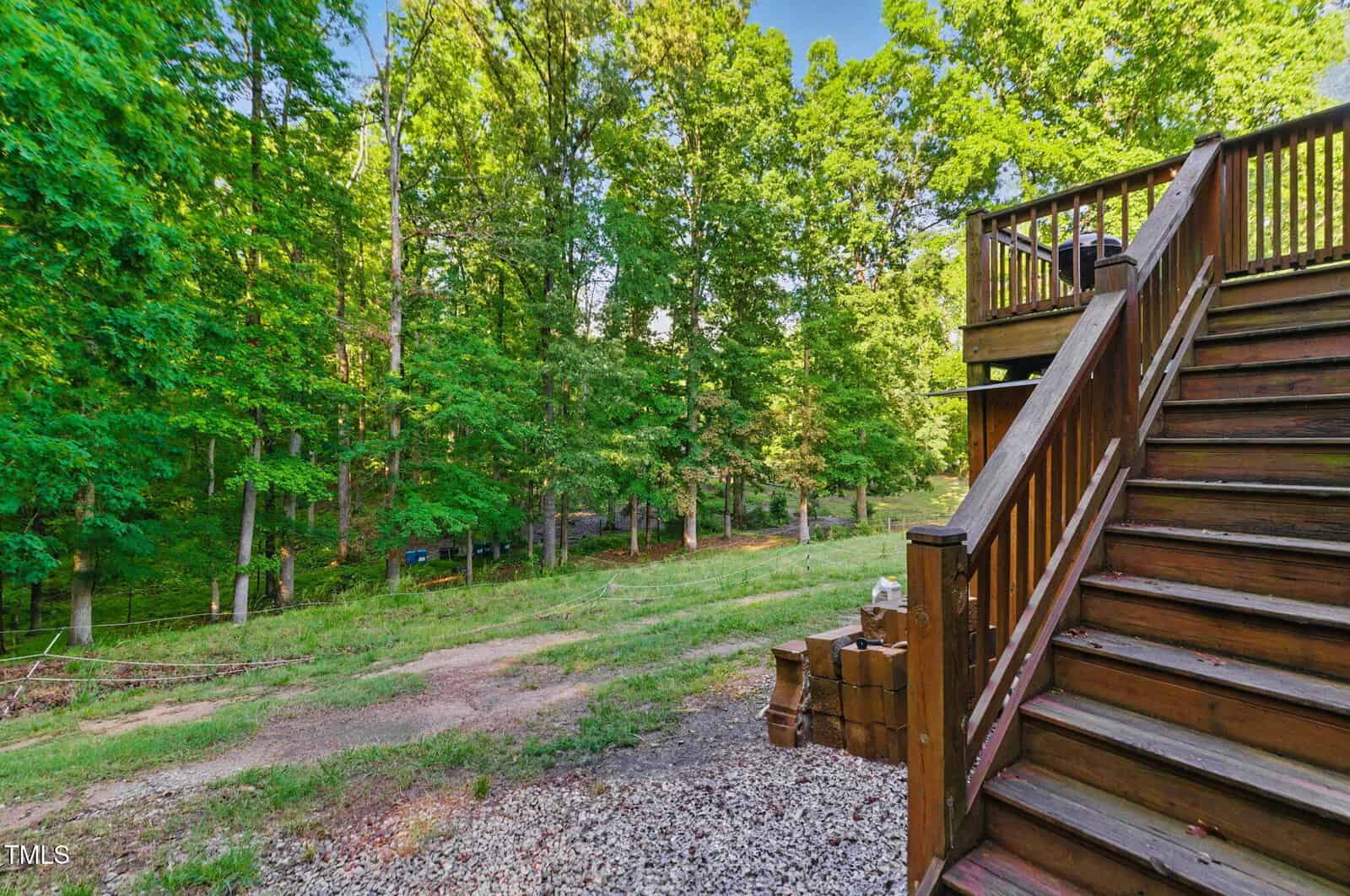 A wooden staircase leads from a deck down to a grassy backyard with scattered rocks and bricks, bordered by a wooded area with dense green trees.