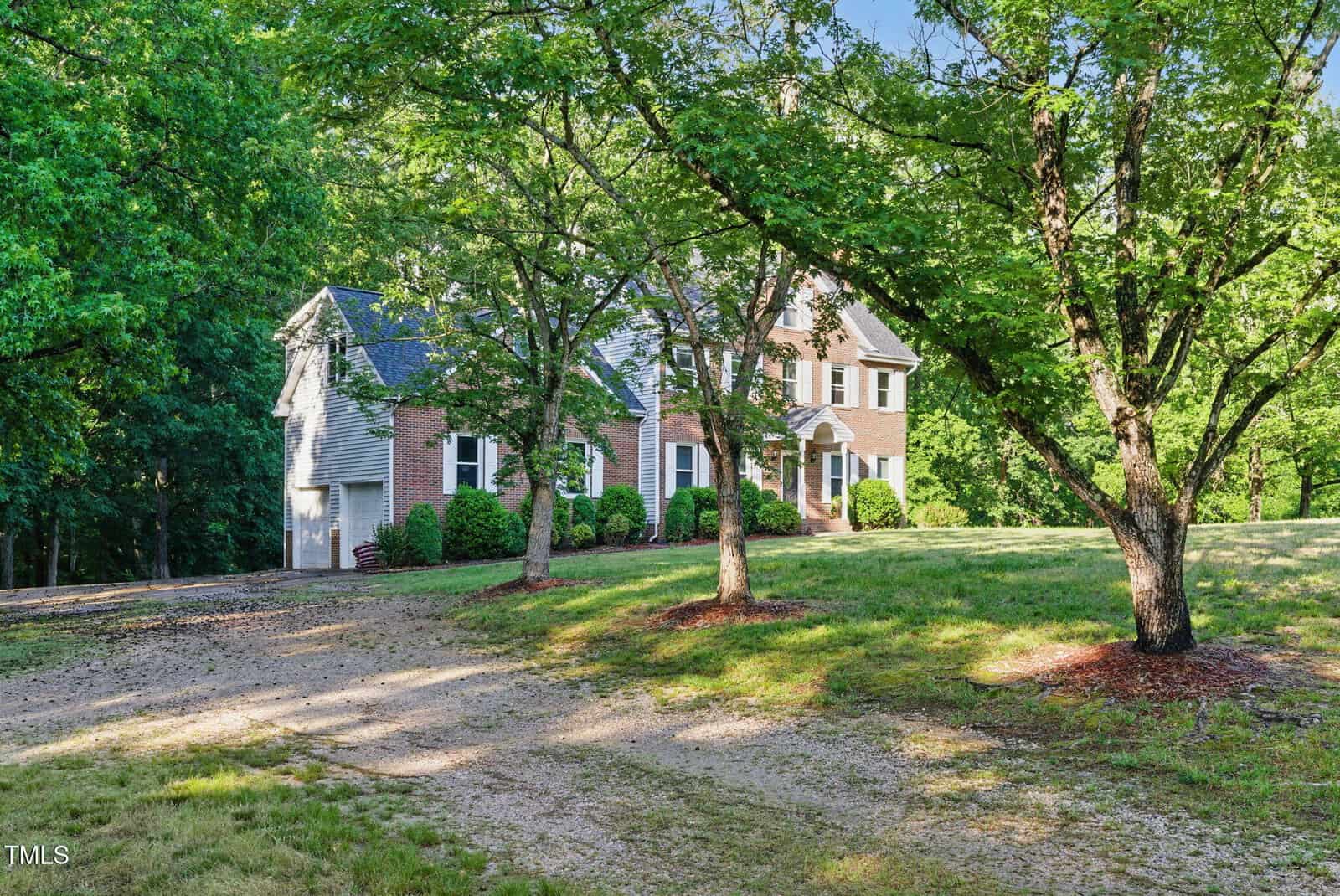 A two-story brick and siding house with white trim, surrounded by green trees and a grassy yard, sits on a spacious lot with a dirt driveway in front. Sunlight filters through the branches, creating a peaceful atmosphere.