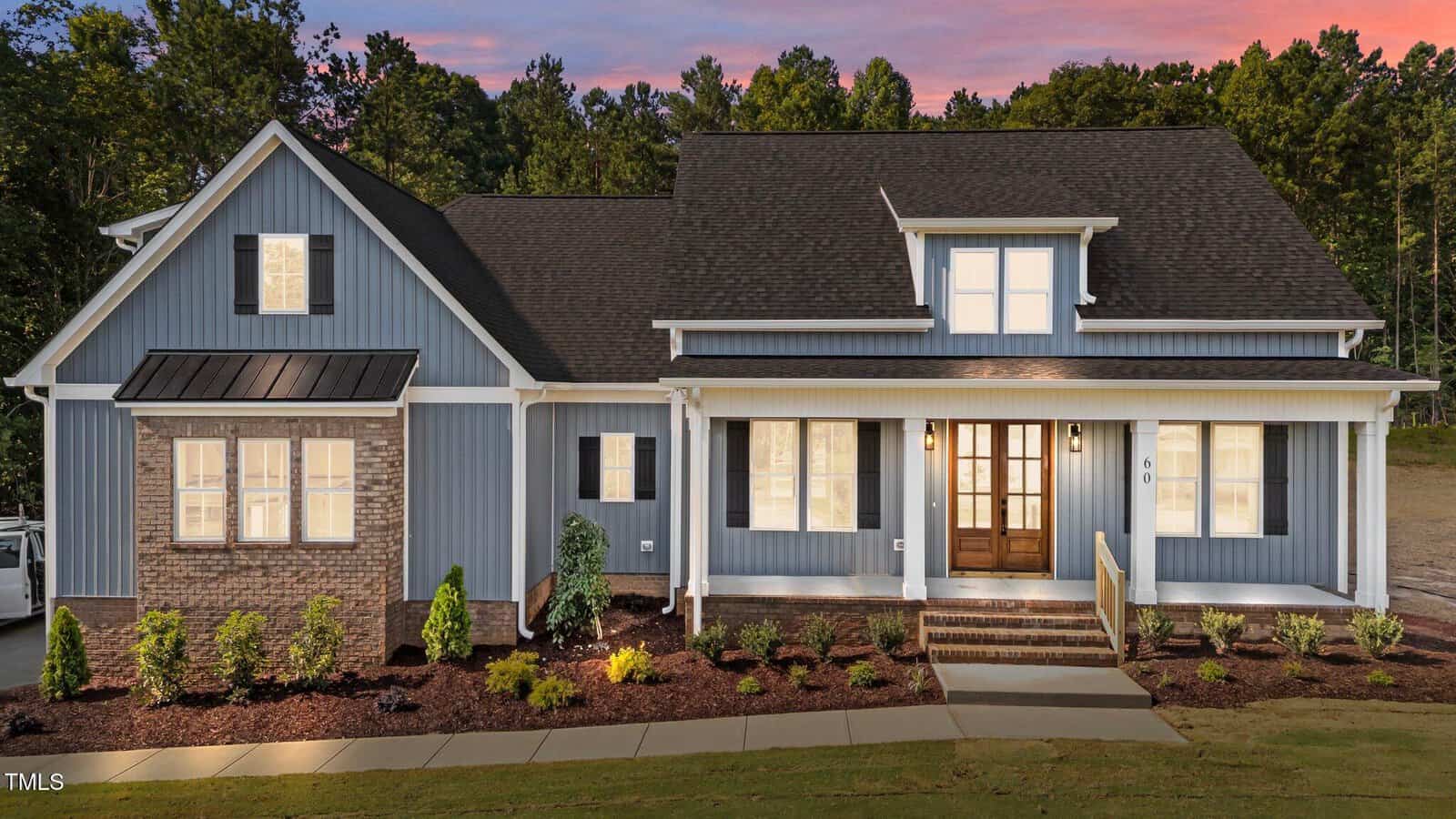 A modern two-story house with blue-gray siding, white trim, a dark shingled roof, and a brick accent. Warmly lit windows and a wooden front door face a landscaped yard at sunset.