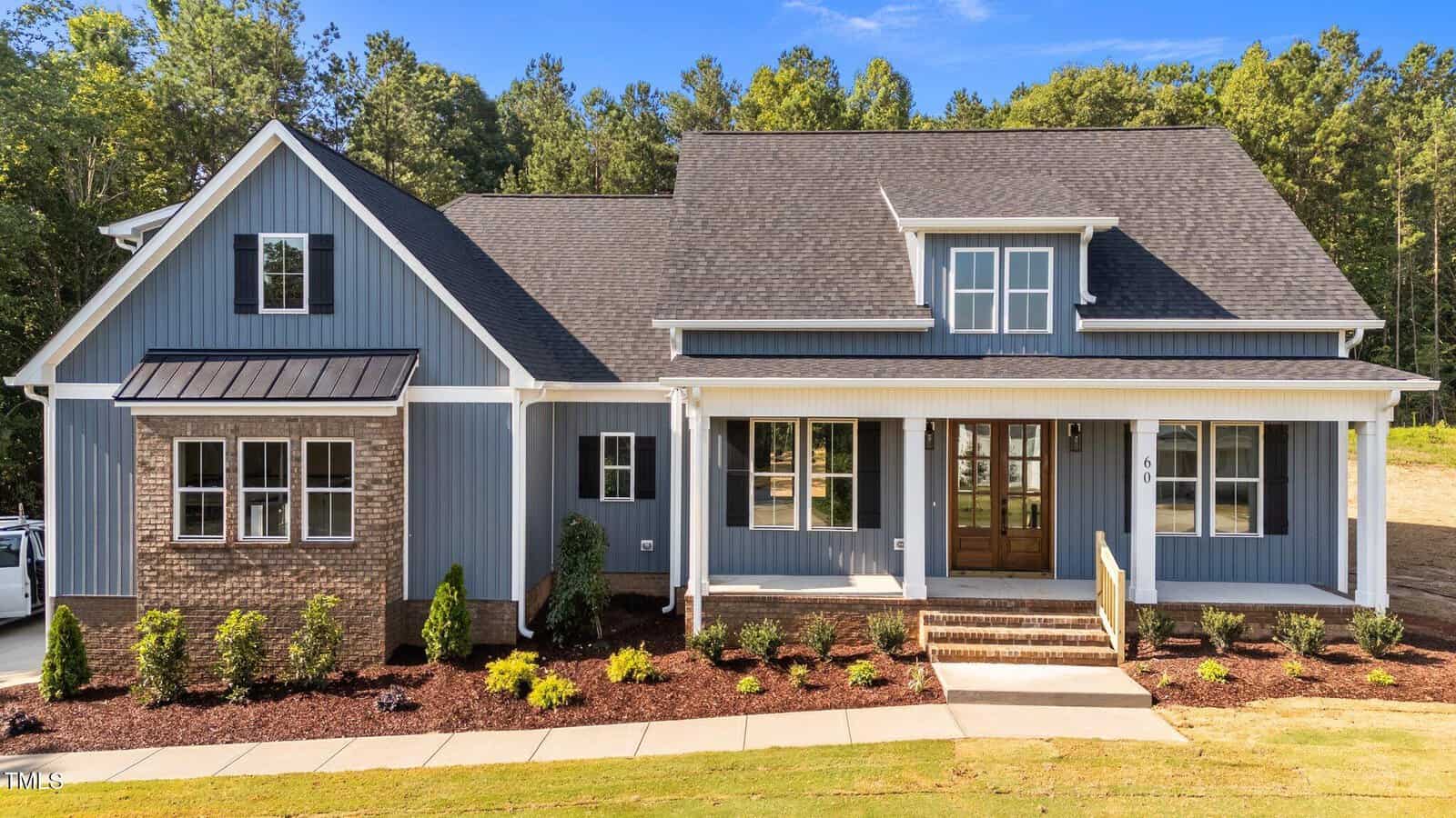 A modern farmhouse-style home with blue-gray siding, white trim, a large front porch, and a wooden double front door. The house has a dark shingle roof, brick accents, and landscaped shrubs in front. Trees are in the background.