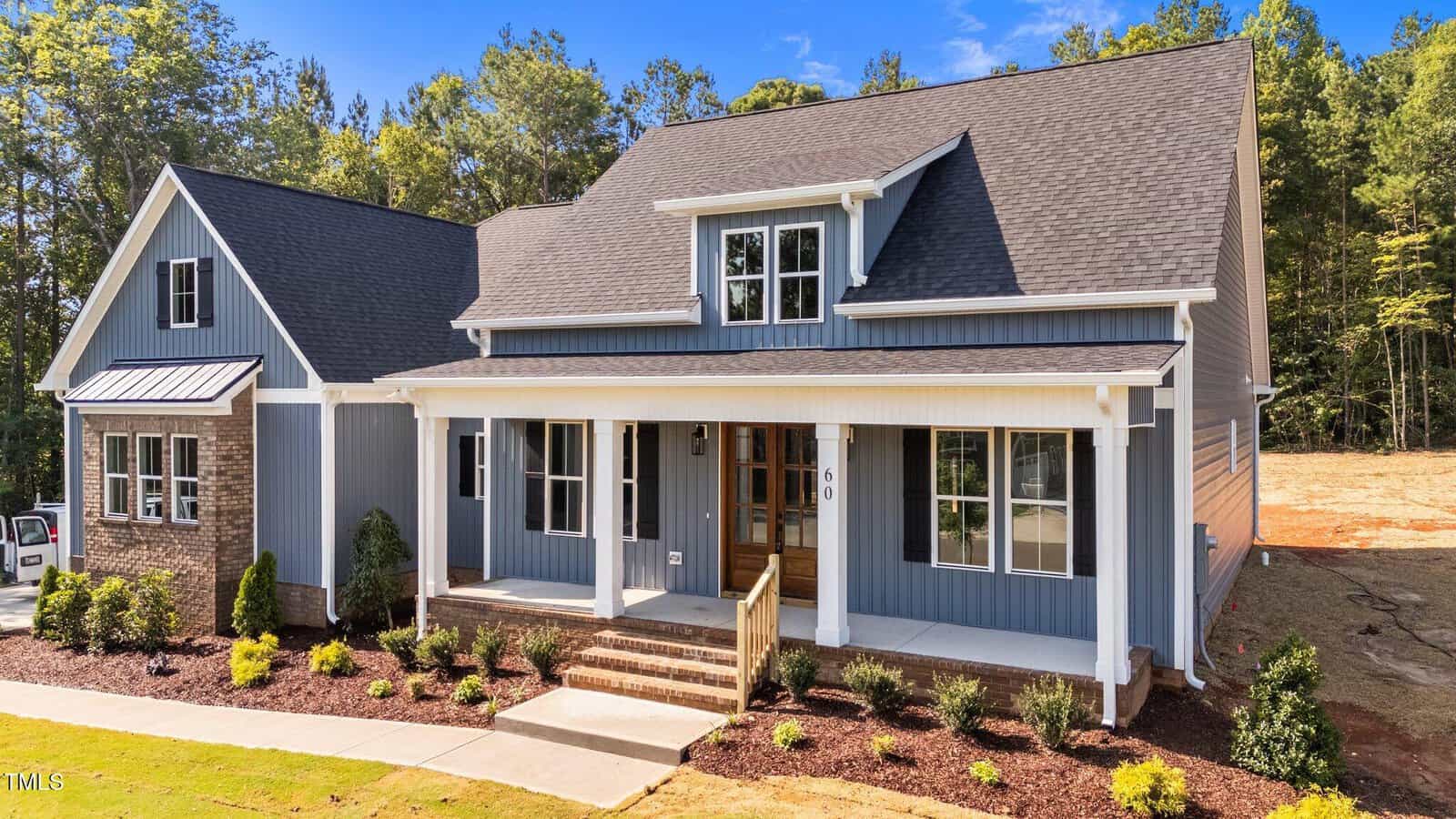 A modern gray and white house with a covered front porch, gabled roof, and brick accents. The yard is landscaped with shrubs and mulch, and trees are visible in the background.
