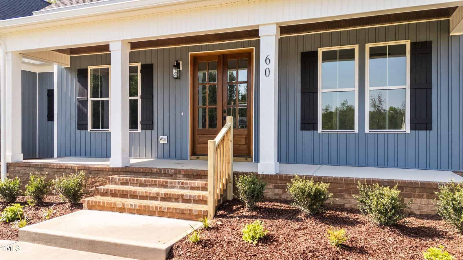 A front porch of a modern house with blue siding, white trim, black shutters, and a wooden door with glass panels. Brick steps lead up to the porch, and small shrubs are planted along the walkway.