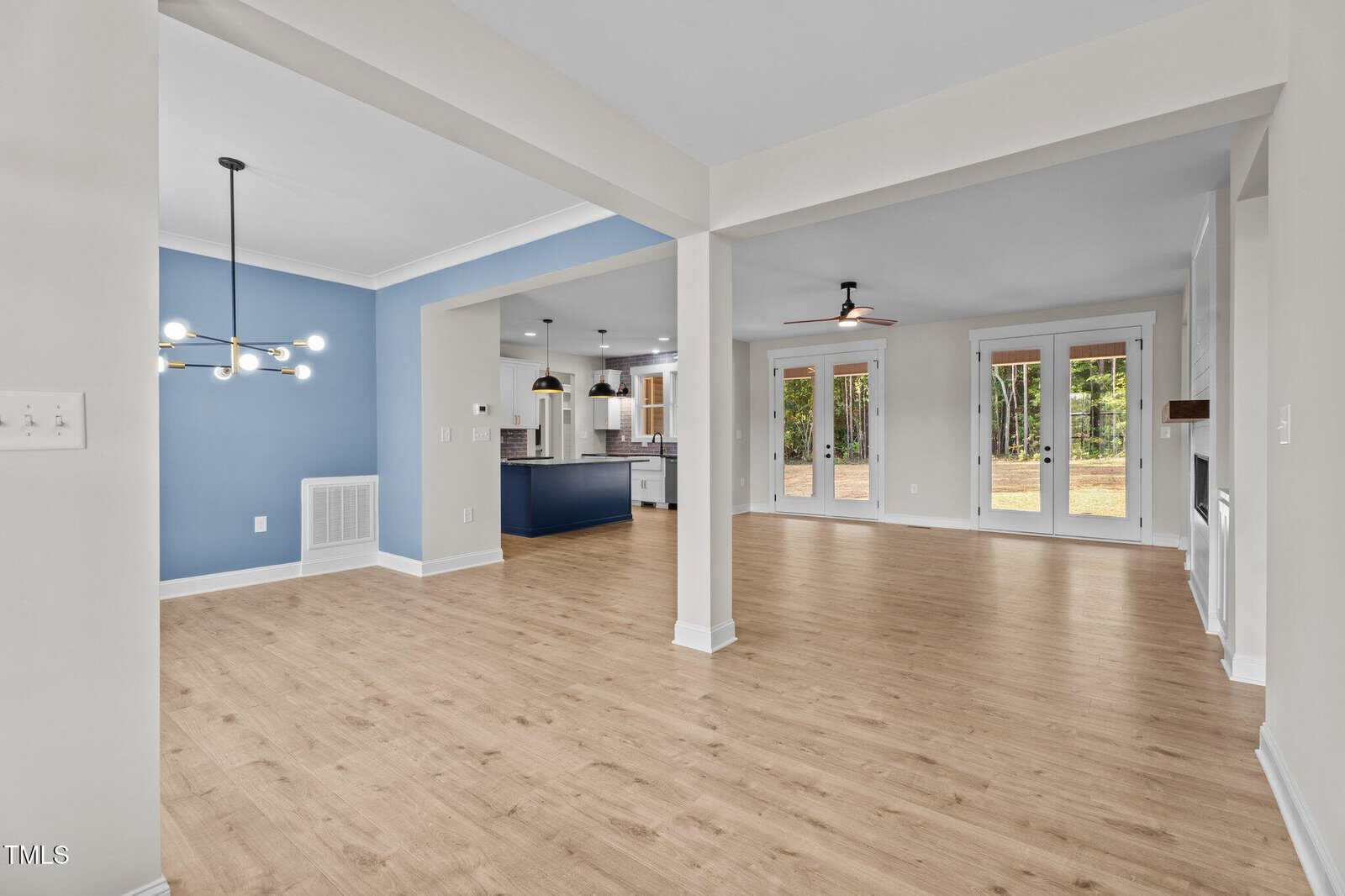 Open-concept living space with light wood floors, a blue accent wall in the dining area with a modern chandelier, and a spacious kitchen with white cabinets, island, pendant lights, and large windows overlooking a backyard.