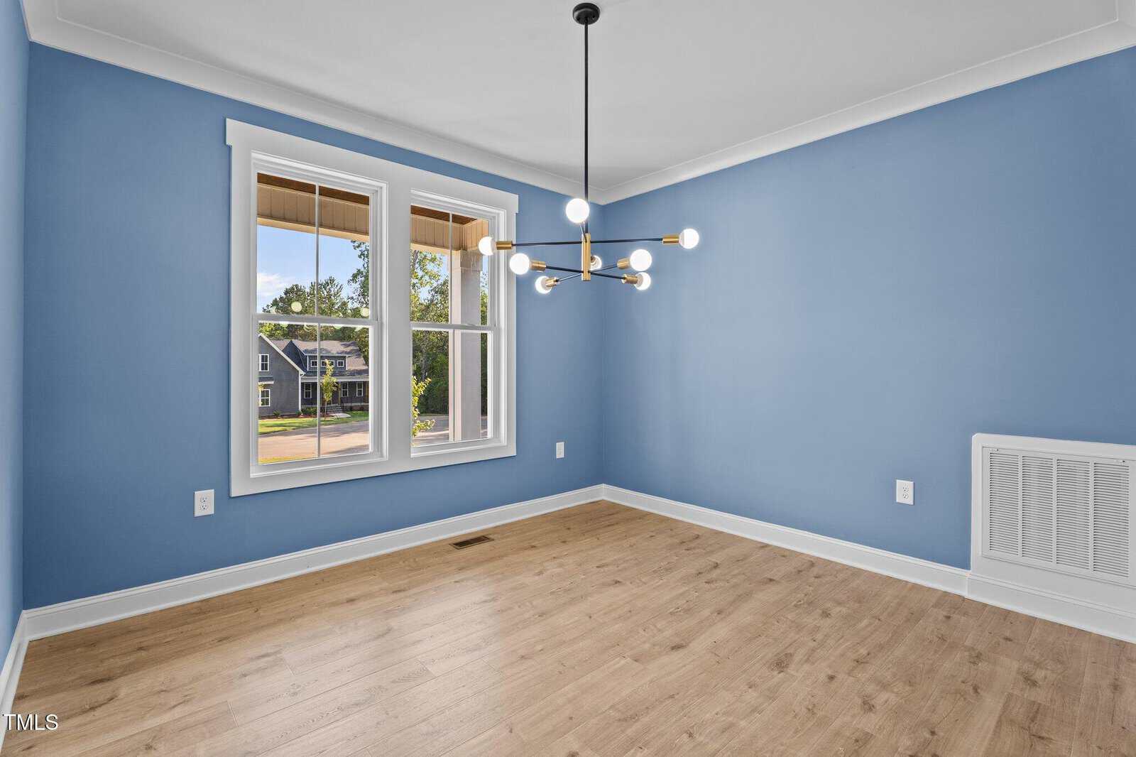 A bright, empty room with light blue walls, wood floor, large window, and a modern black and gold chandelier hanging from the ceiling.