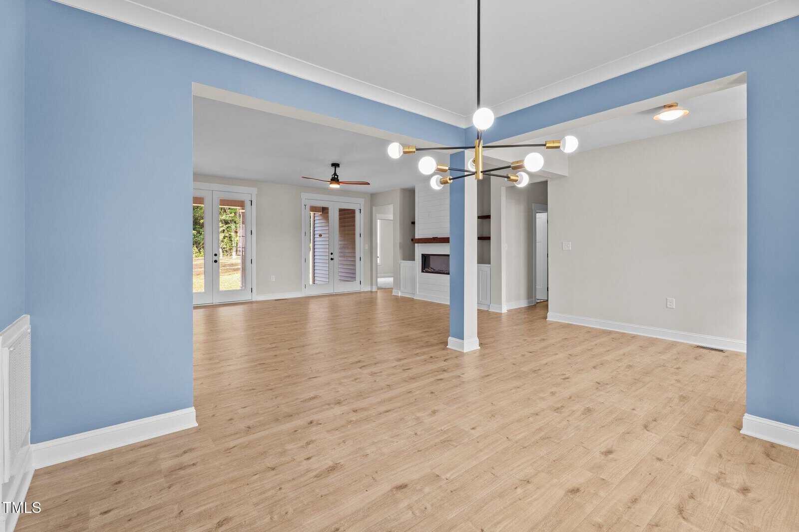 Open-concept living and dining area with light wood floors, blue and white walls, a modern chandelier, and large windows letting in natural light. A fireplace is visible in the adjacent room.