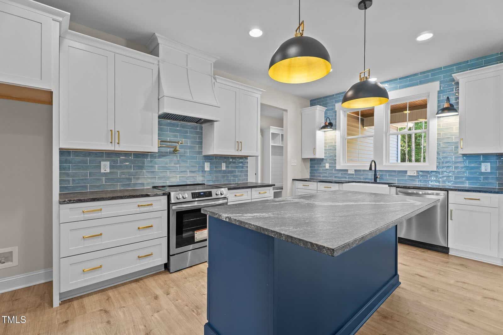 Modern kitchen with white cabinets, a blue island, gold hardware, gray countertops, stainless steel appliances, blue subway tile backsplash, wood floors, pendant and recessed lighting, and a window over the sink.