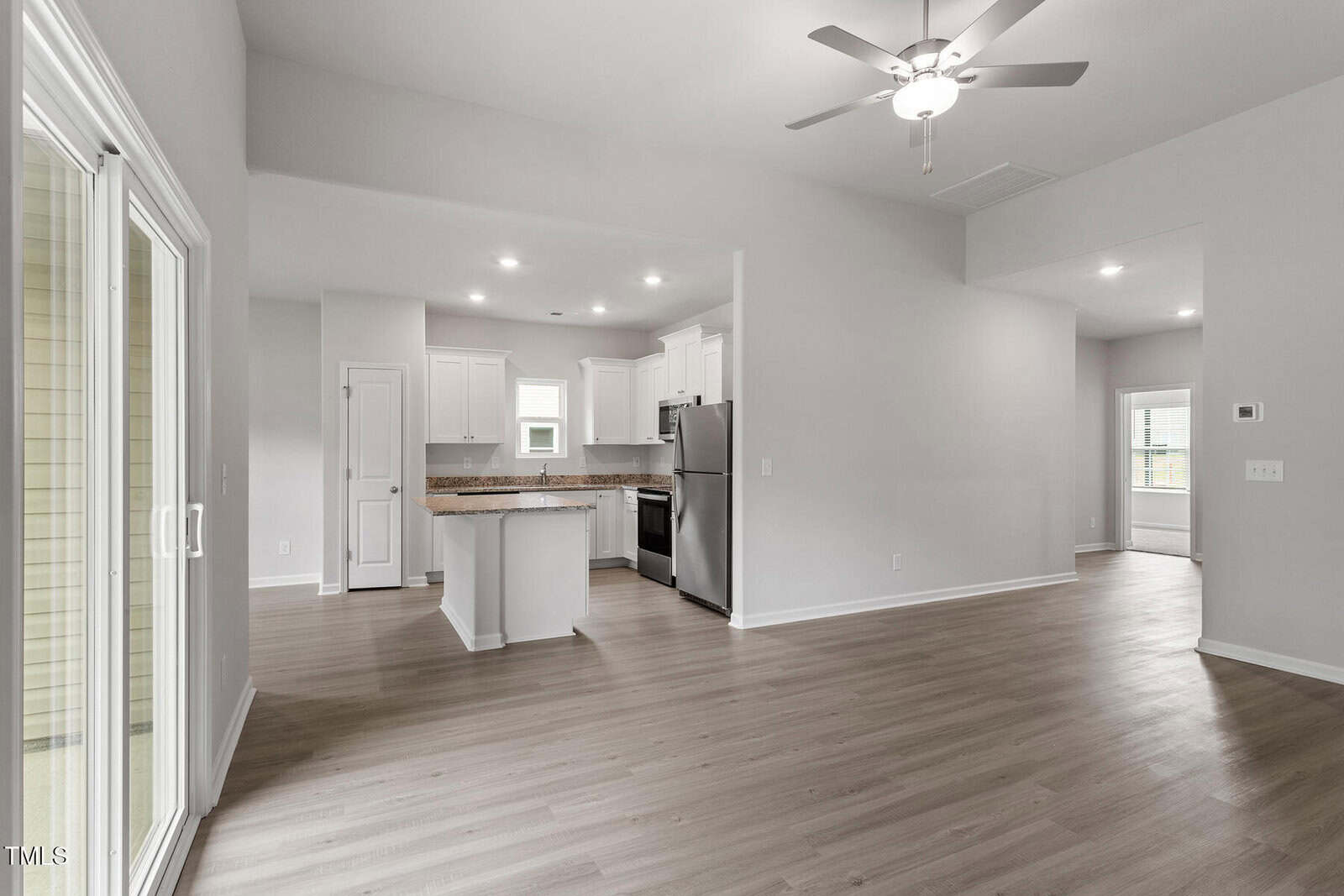 A modern, unfurnished open-plan living area with light wood flooring, white walls, a ceiling fan, and a kitchen featuring stainless steel appliances and recessed lighting. Sliding glass doors are visible on the left.