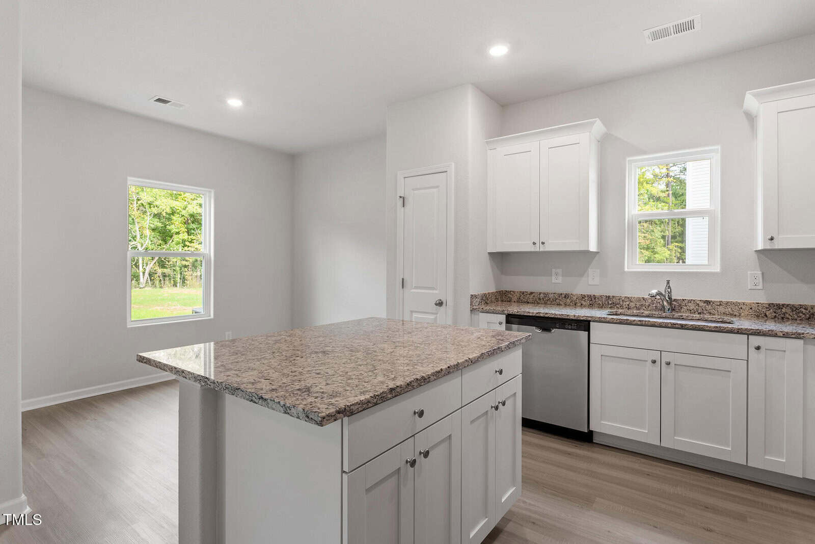 Bright kitchen with white cabinets, light-colored granite countertops, an island, stainless steel dishwasher, and two windows. The space features light wood flooring and neutral wall colors.
