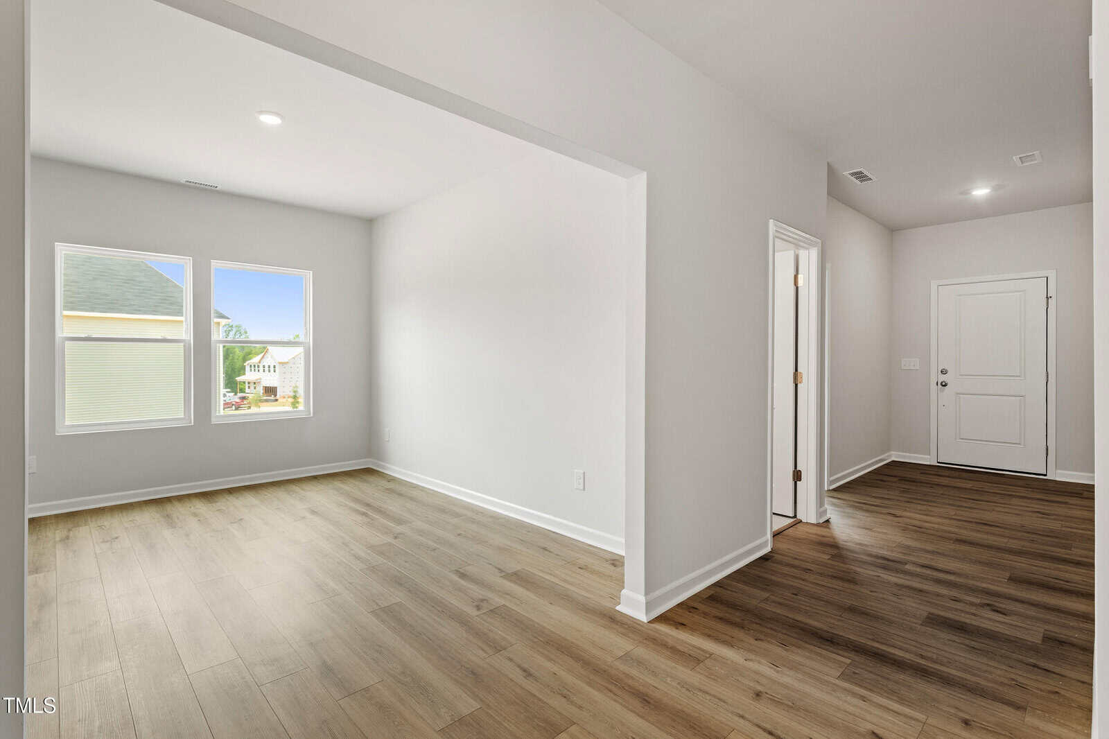 A bright, empty room with light-colored walls and wood flooring, featuring a window with natural light and an open doorway leading to a hallway with a white front door.