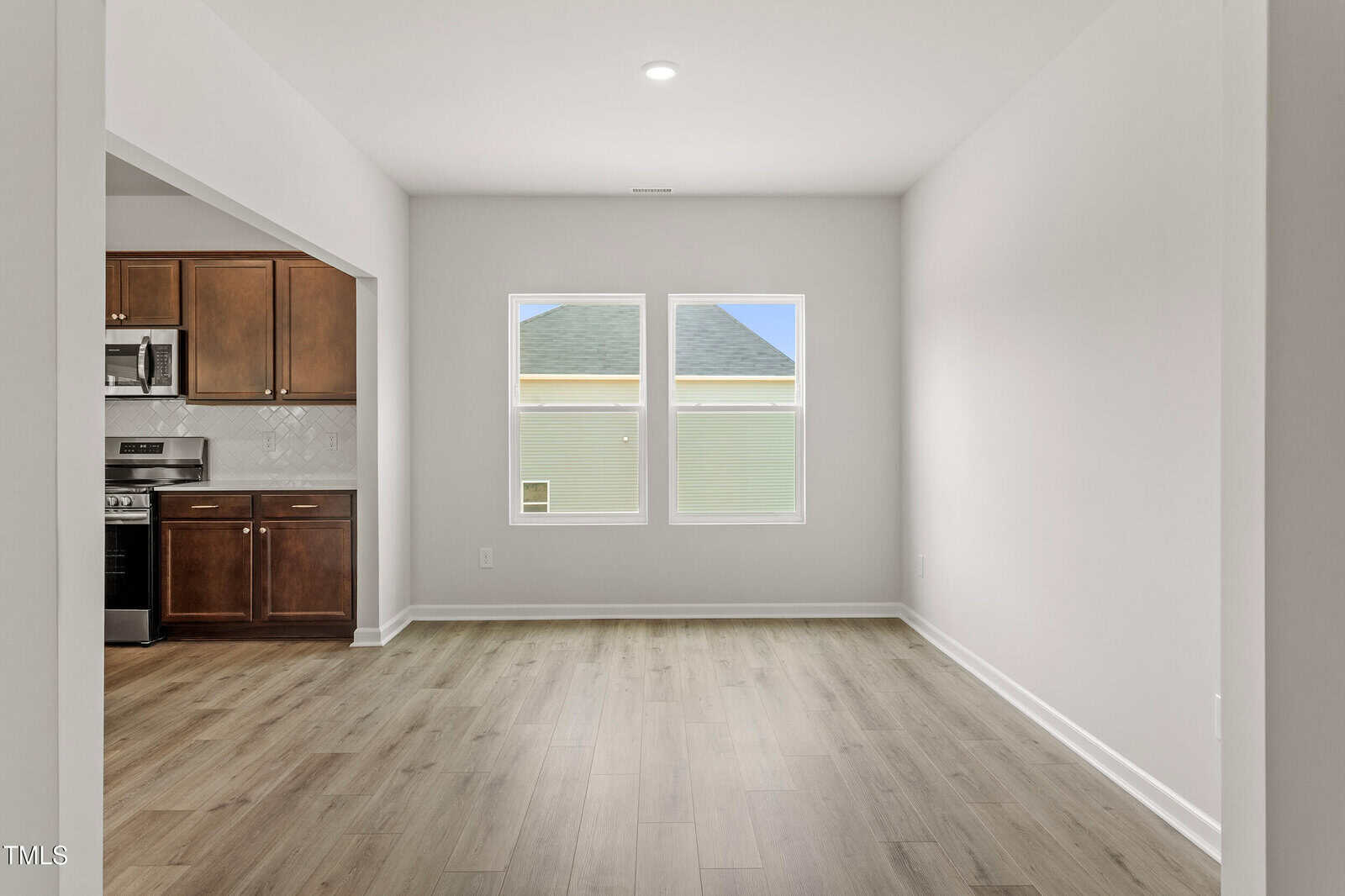 A bright, empty room with light wood flooring, white walls, and two large windows. An adjacent kitchen with brown cabinets and stainless steel appliances is visible on the left.