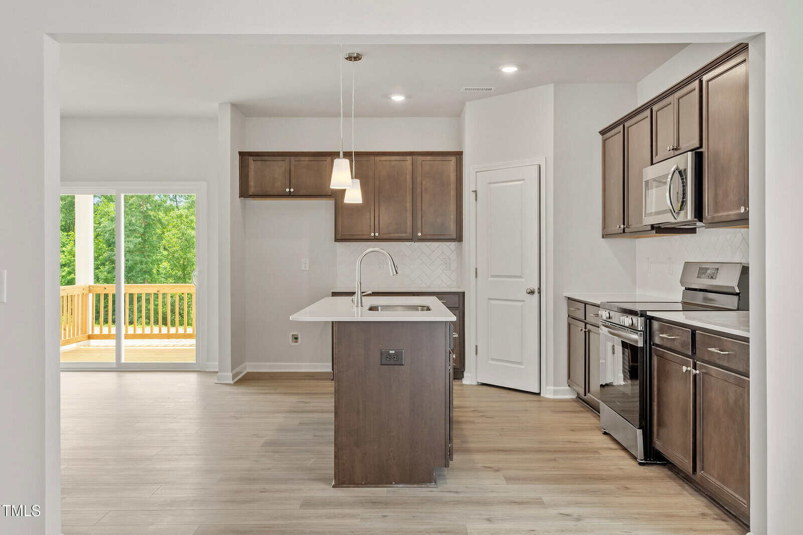 Modern kitchen with dark wood cabinets, stainless steel appliances, an island with pendant lights, and light wood flooring. Sliding doors lead to a wooden deck with a view of green trees outside.