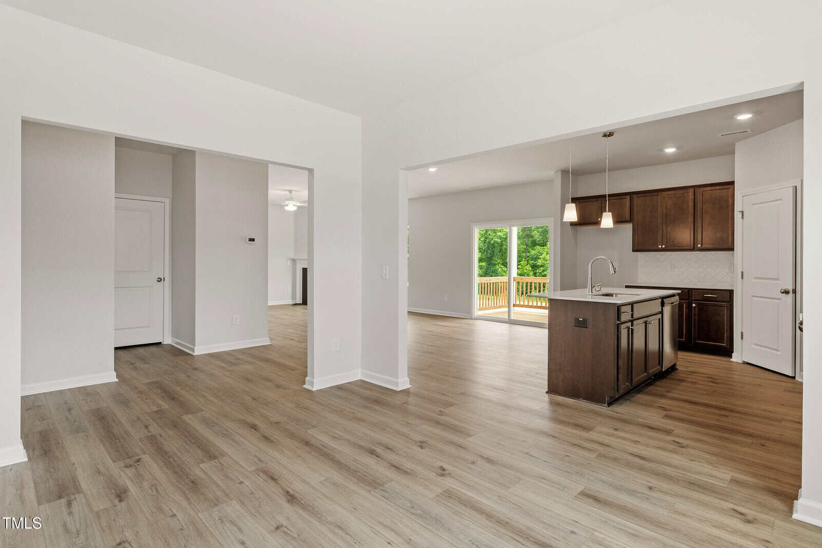 Open-concept living space with light wood flooring, white walls, and a modern kitchen with dark cabinets and an island. Large sliding glass doors lead to a wooden deck with greenery outside.