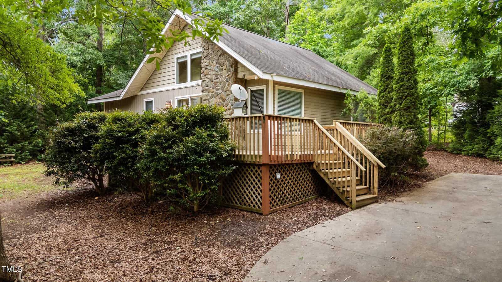 A small tan cottage with a stone chimney and wooden porch sits surrounded by trees and bushes. A short staircase leads to the raised entryway, and a concrete path curves in front of the house.
