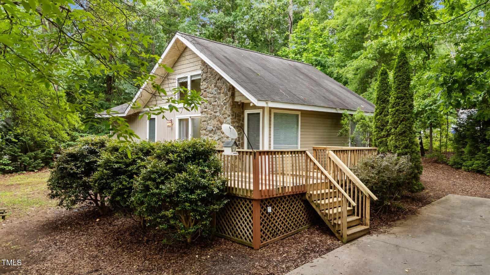 A small house with stone and siding exterior, surrounded by trees and bushes, features a wooden porch with railing and stairs leading to the entrance. The house sits on a mulched yard next to a concrete driveway.