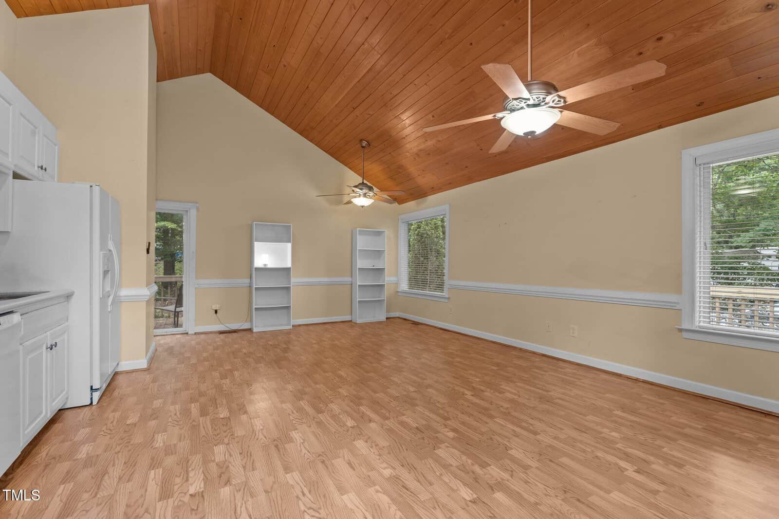 Spacious room with wood laminate flooring, vaulted wood-paneled ceiling, two ceiling fans, large windows, white kitchen cabinets on the left, two white bookshelves, and a glass door leading outside.