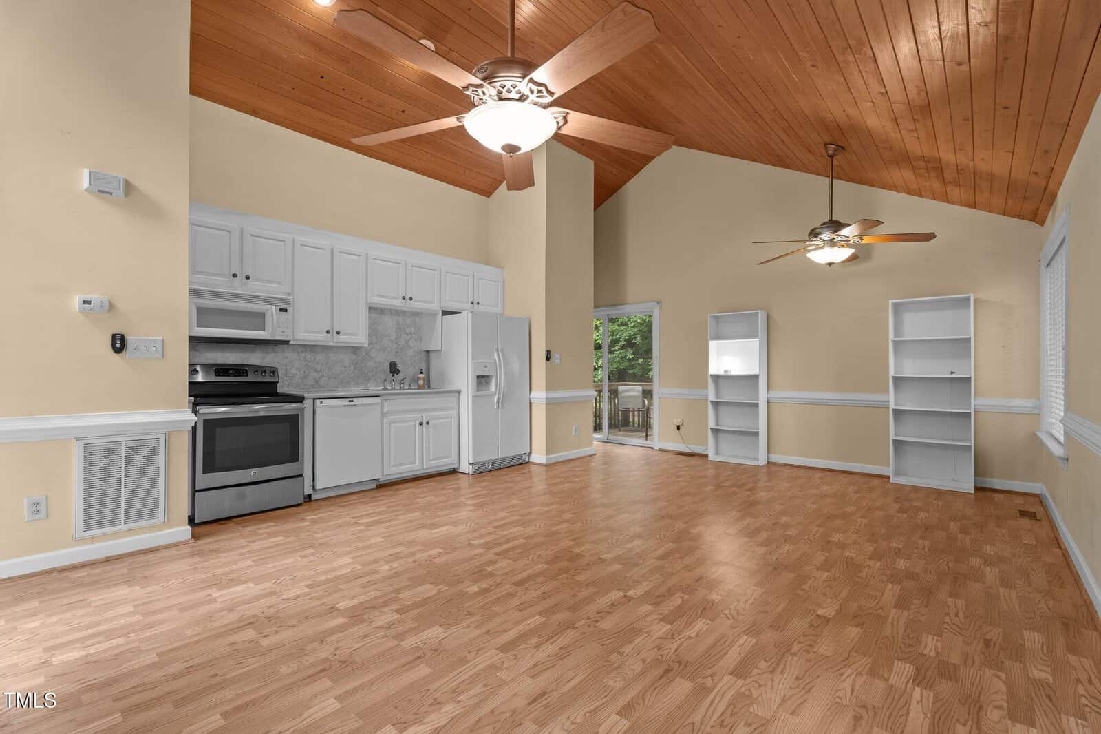 Open-plan room with a wood ceiling, ceiling fans, light wood floors, white kitchen cabinets, stainless steel appliances, and two white bookshelves near sliding glass doors leading outside. Walls are painted a soft beige color.