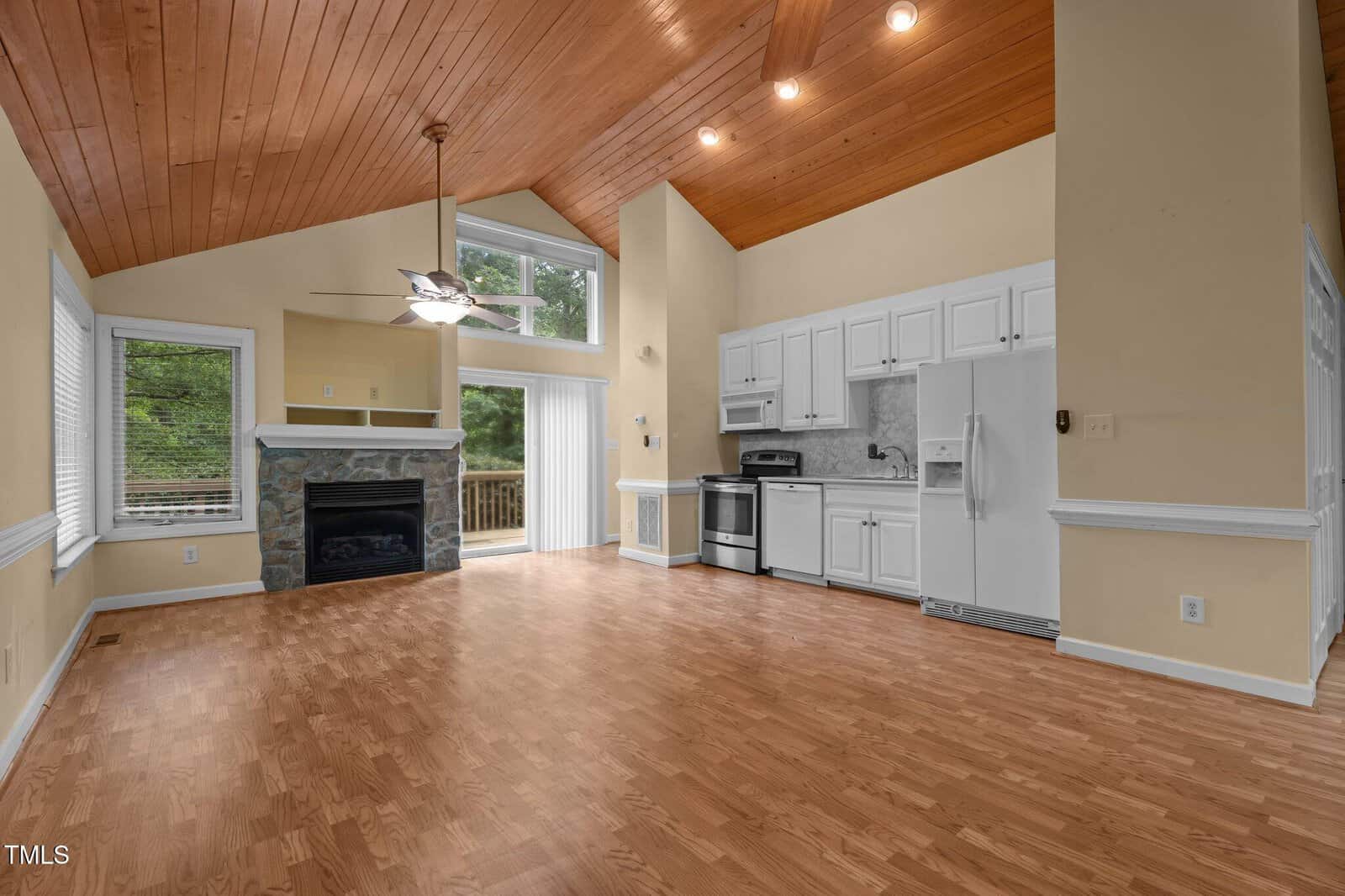 Open-concept room with wood floors and a vaulted wooden ceiling, featuring a kitchen with white cabinets and appliances, a stone fireplace, large windows, and sliding glass doors leading outside.