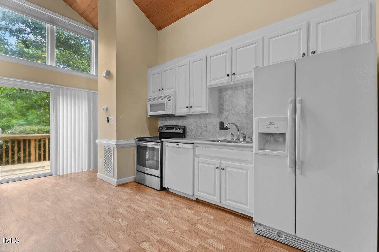 Modern kitchen with white cabinets, stainless steel stove, microwave, dishwasher, and white refrigerator. Light wood flooring and large windows with a view of trees and a wooden deck outside. High ceilings with wood accents.