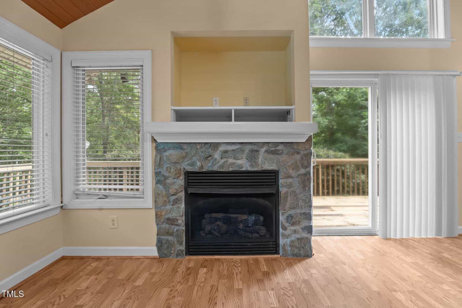 A stone fireplace with a black insert is centered between two windows and in front of a glass door leading to a wooden deck. The room has light wood floors and light-colored walls. Some trees are visible outside.