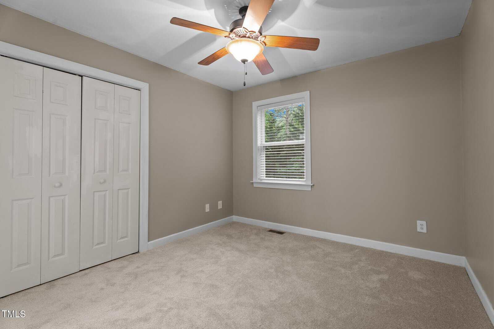 Empty carpeted room with beige walls, white baseboards, a window with white blinds, ceiling fan with light, and double white closet doors. Electrical outlets are visible on the walls.