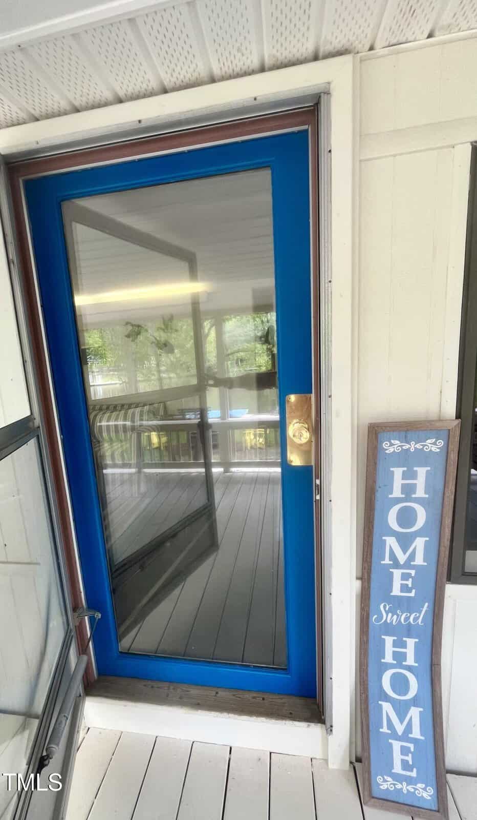 A blue storm door with a gold handle is set in a white wall. A vertical sign next to the door reads “Home Sweet Home” in white letters on a blue background. The porch is covered and enclosed with windows.