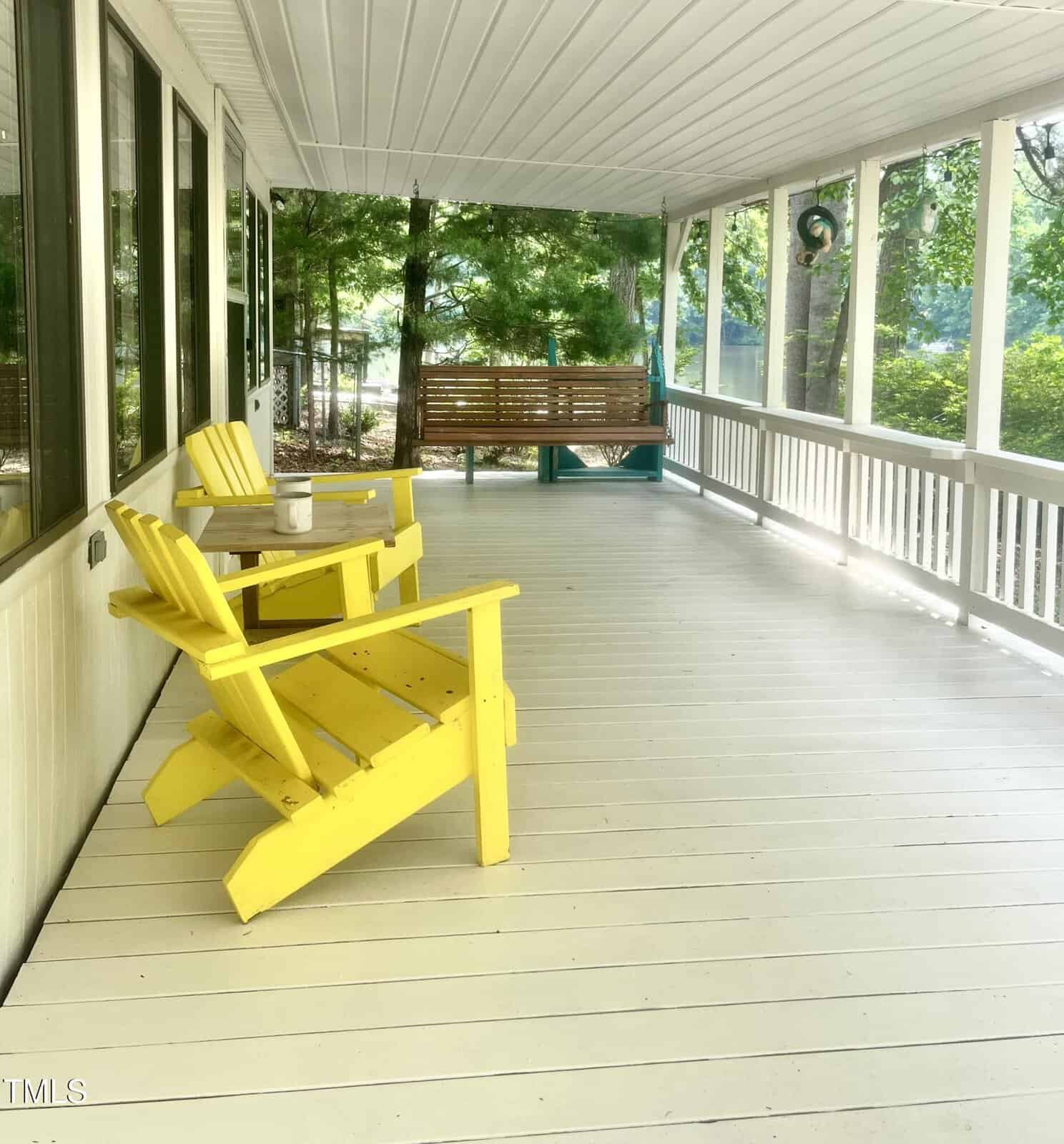 A spacious white porch with two yellow Adirondack chairs, a small side table holding a mug, and a wooden porch swing. The porch is surrounded by trees and partially shaded.