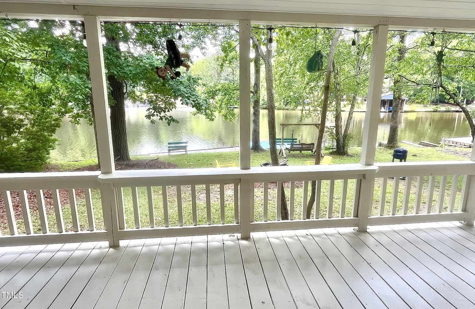 View from a white wooden porch overlooking a grassy yard with trees, a lake, benches, a swing, Adirondack chairs, and a small dock in the distance. The scene is bright and the area appears peaceful.