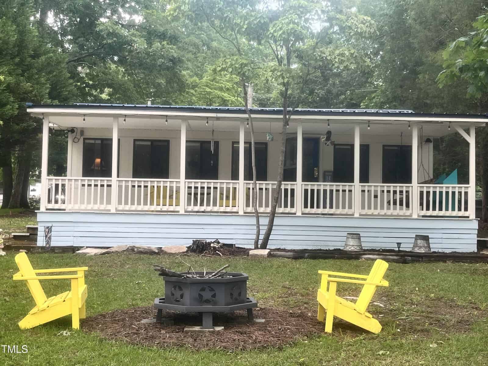 A cozy light blue house with a wide front porch and white railing sits in a green yard. Three bright yellow Adirondack chairs surround a fire pit in front of the house, with trees and string lights in the background.