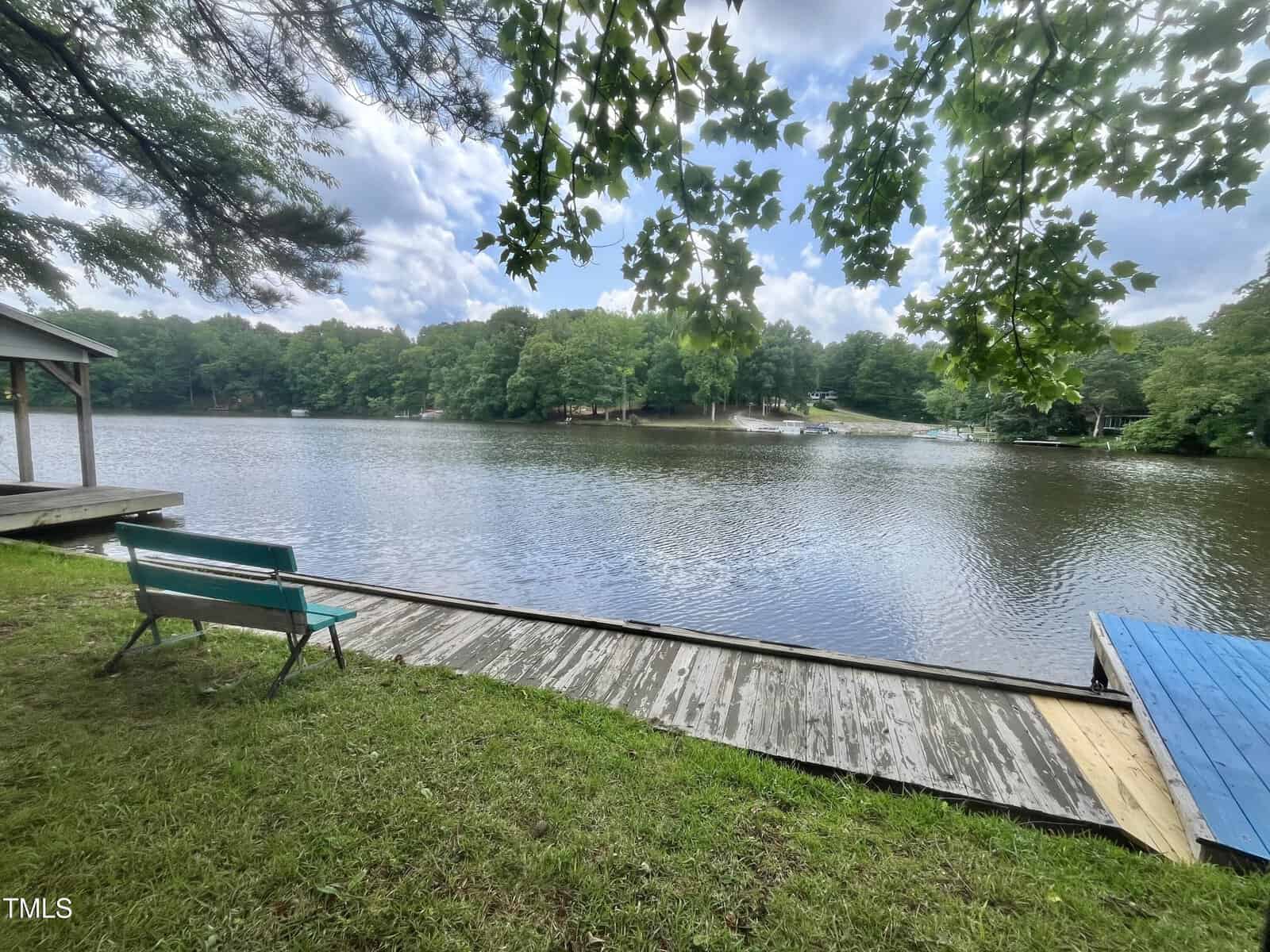 A green bench sits on grass by a wooden dock, overlooking a calm lake surrounded by trees under a partly cloudy sky. Leaves hang overhead, and a covered structure is visible to the left.
