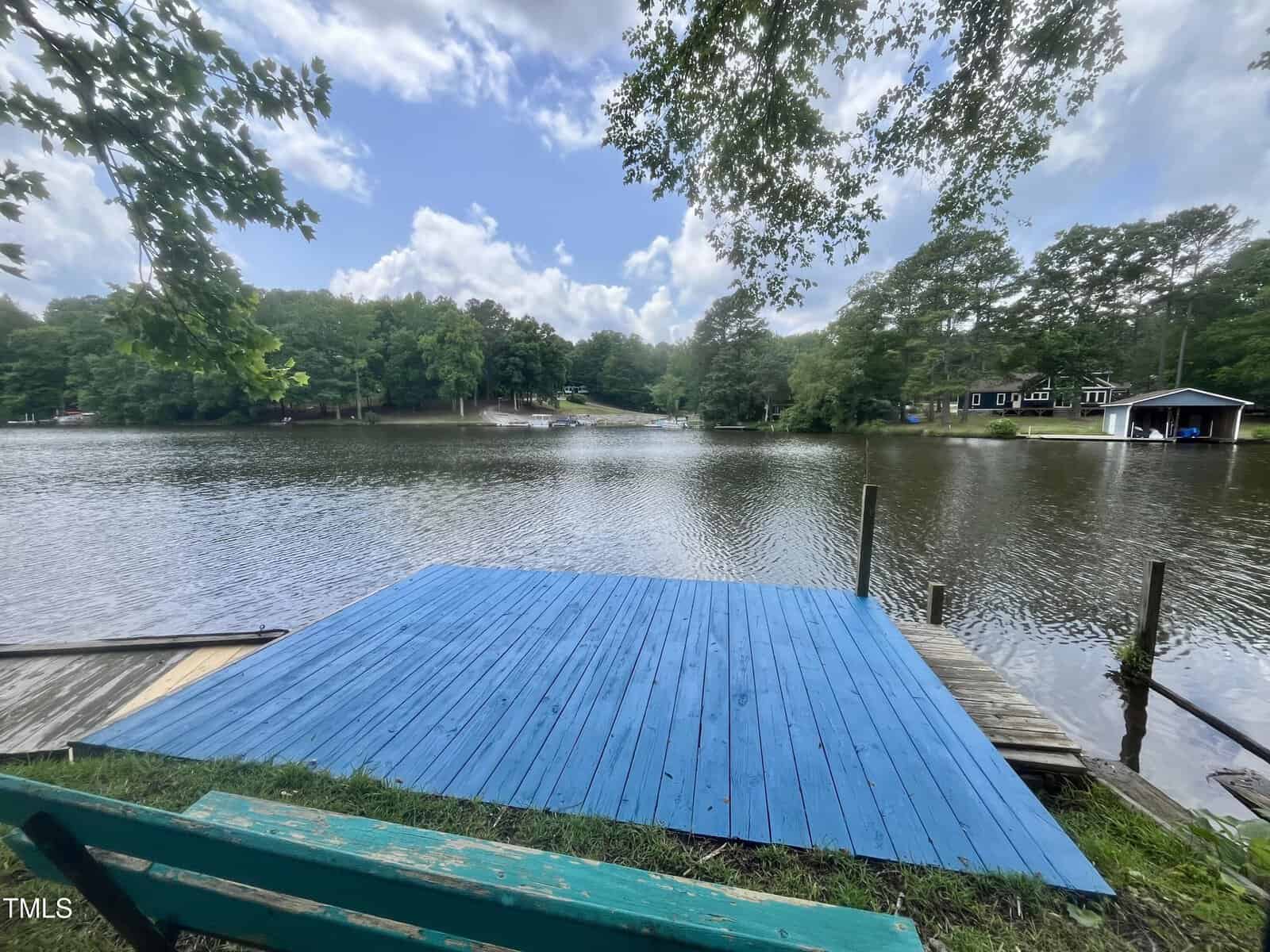 A bright blue wooden dock extends over a calm lake, surrounded by green trees and houses. The sky is partly cloudy, and a green bench sits in the foreground facing the water.