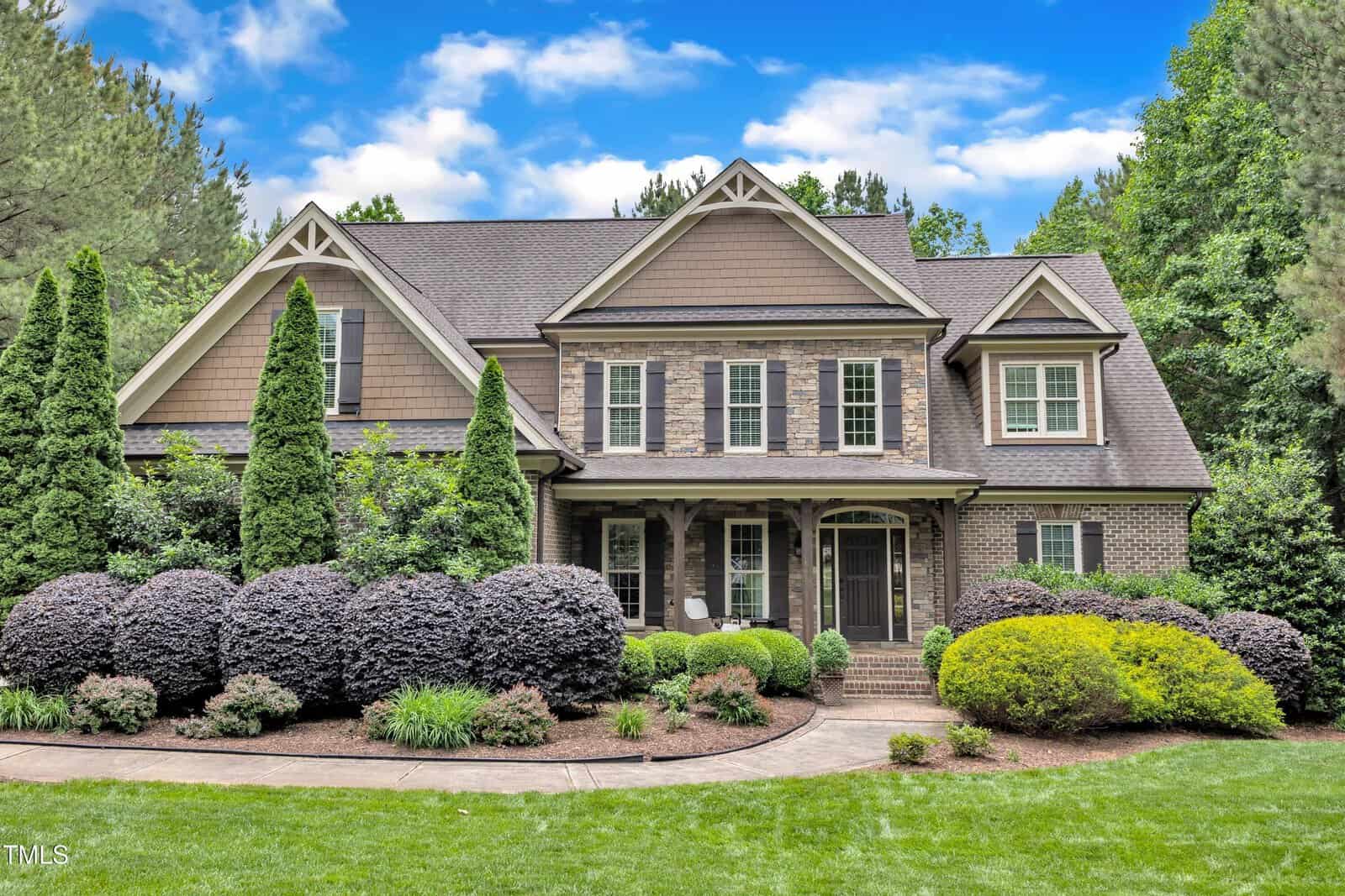This large two-story house at 3007 Krogen Court in Creedmoor features a brick and siding exterior, multiple gables, a covered front porch, and well-maintained landscaping with bushes, shrubs, and trees under an overcast sky with scattered clouds.