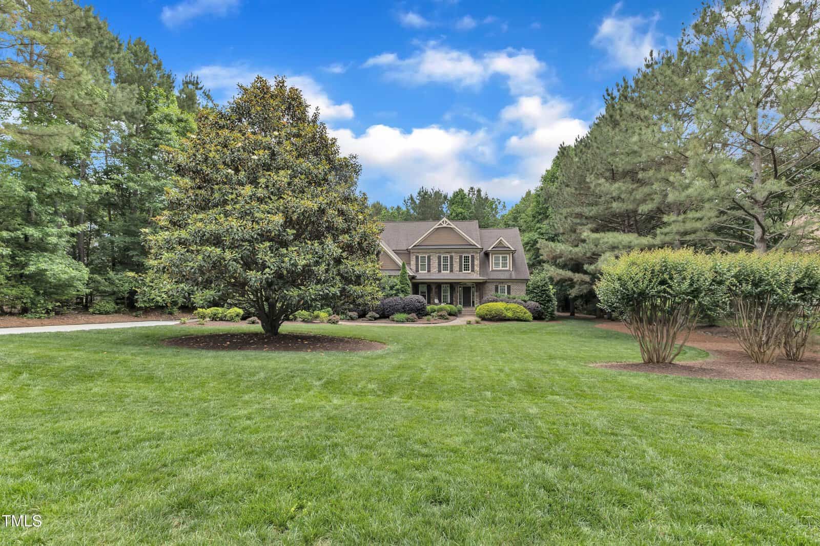 A two-story house with beige siding and large windows is set back from a well-manicured lawn, surrounded by mature trees and landscaped shrubs under a blue sky with scattered clouds.