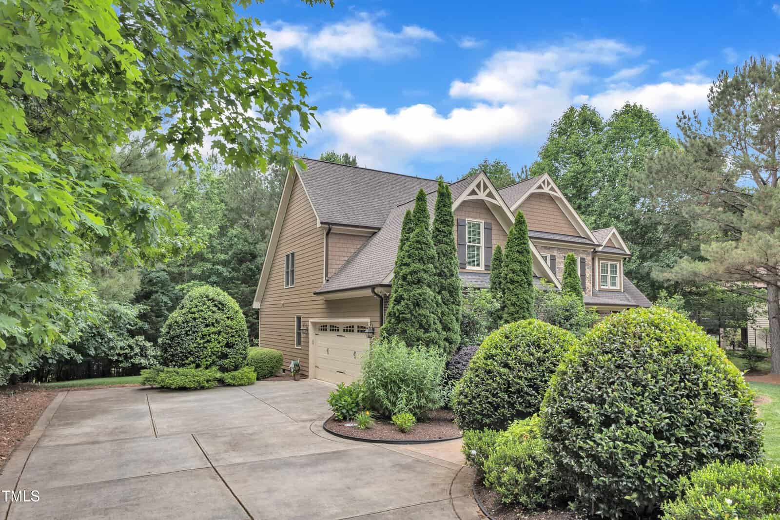 A two-story beige house with a double garage sits at the end of a wide concrete driveway, surrounded by manicured shrubs, tall trees, and lush greenery under a partly cloudy sky.