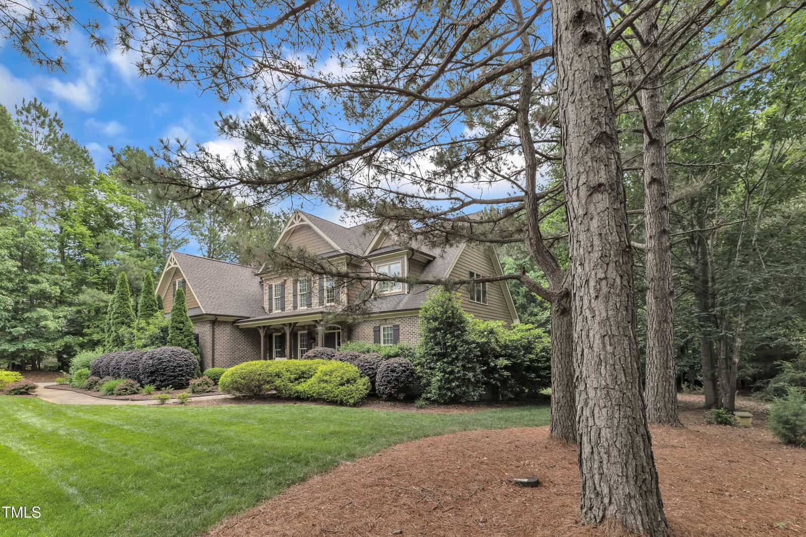 Two-story house with brick and siding exterior surrounded by trees and landscaped bushes. The lawn is well-maintained, and tall pine trees are visible in the foreground under a partly cloudy sky.