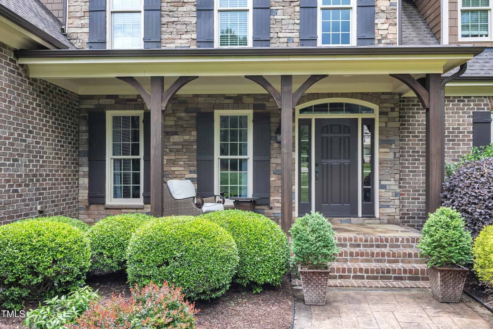 A brick and stone house with a covered front porch, dark front door, two windows, a wicker chair with cushion, and trimmed bushes and shrubs in the landscaped yard.