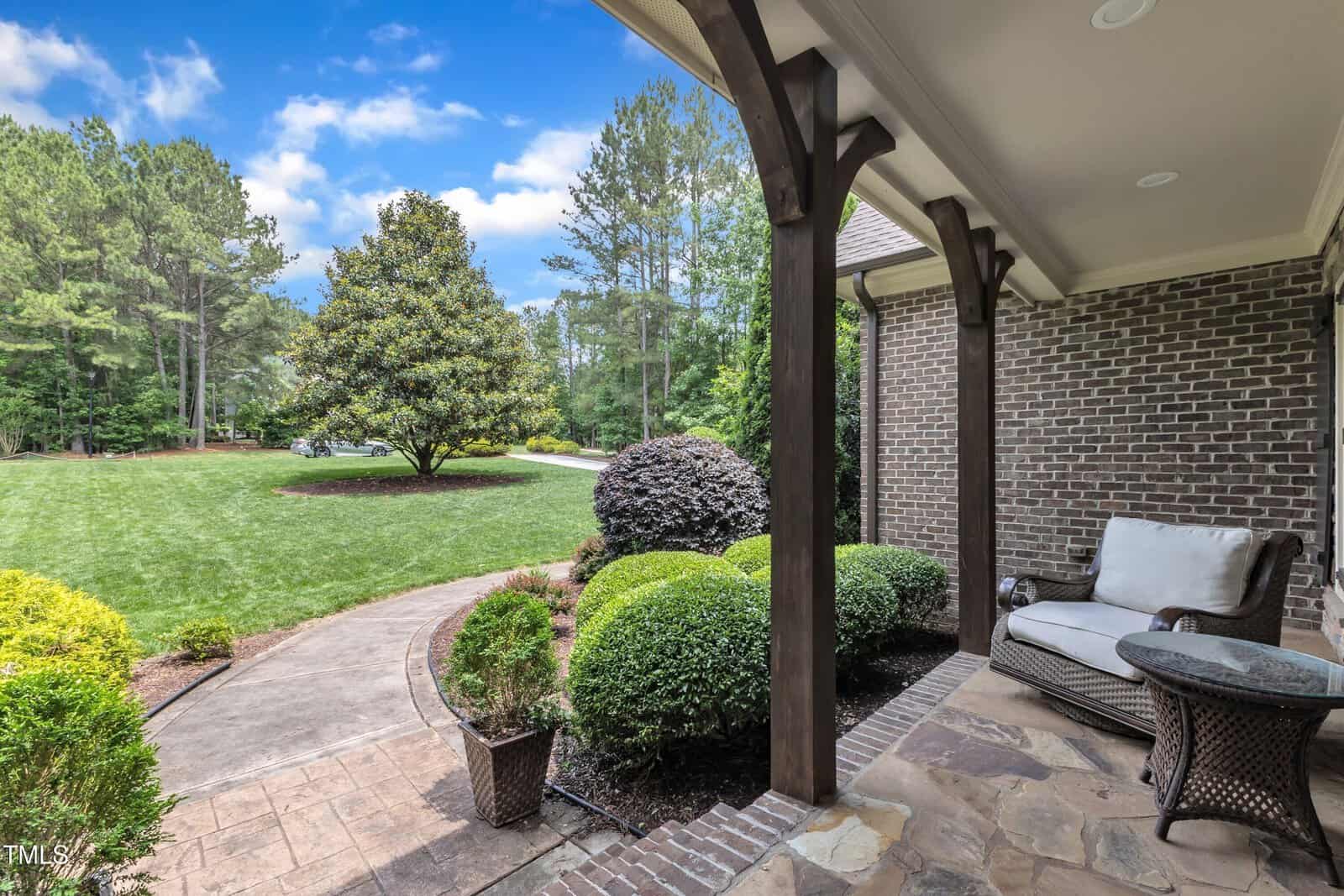 View from a covered patio showing a cushioned chair, a small table, well-maintained shrubs, and a walkway leading to a large, open grassy yard bordered by trees under a partly cloudy sky.