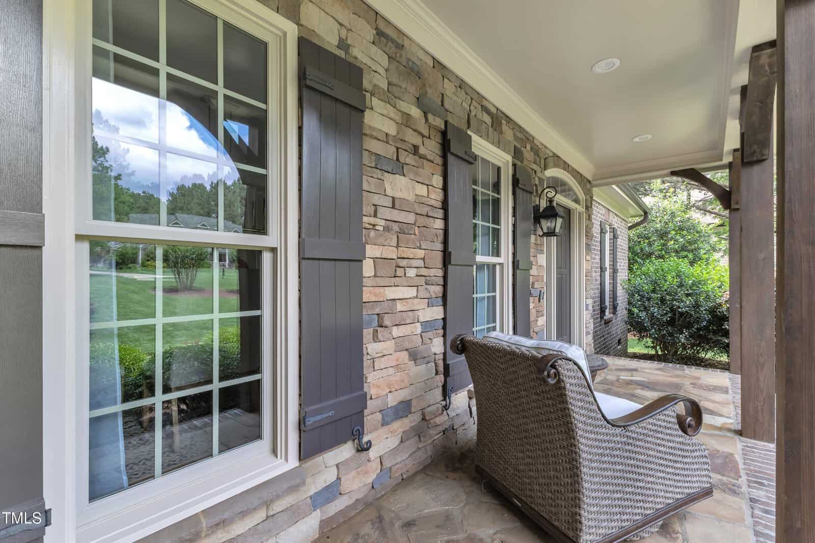 A wicker chair sits on a stone porch outside a house with stone and brick walls, shuttered windows, and a covered ceiling. Bushes and a grassy yard are visible in the background.